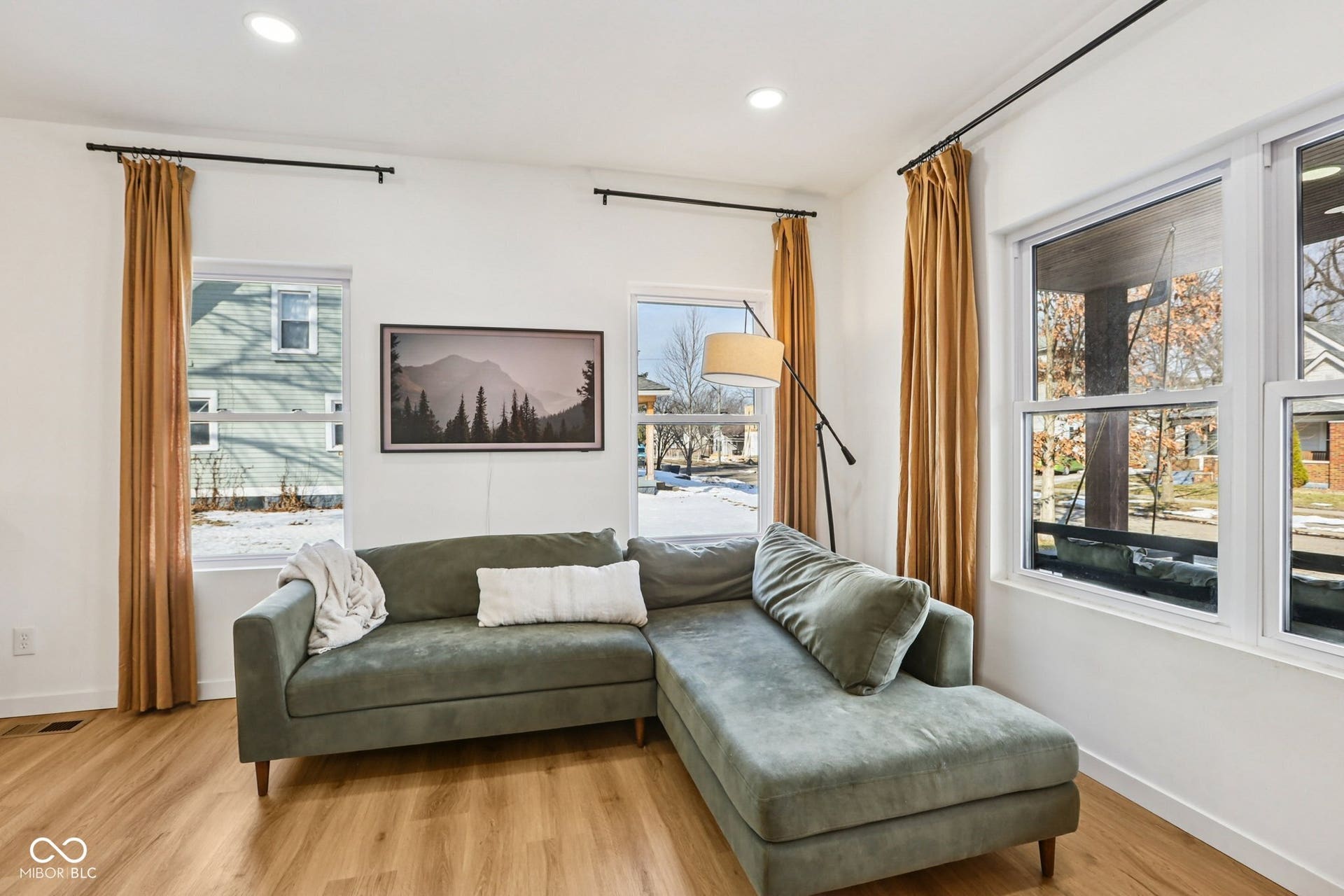Living area featuring wood-finish flooring, white walls, multiple windows with white frames, recessed ceiling lighting, and black curtain rods