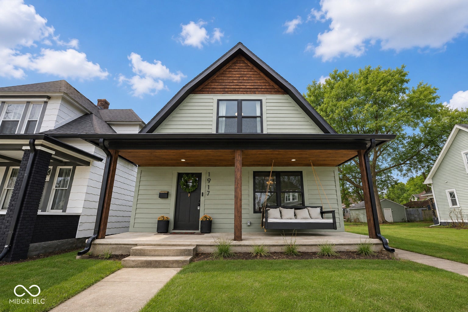 Virtually Altered - Craftsman-style home featuring light green siding, a prominent dark gray front door with transom window, and a covered front porch with exposed wood ceiling and dark wood columns