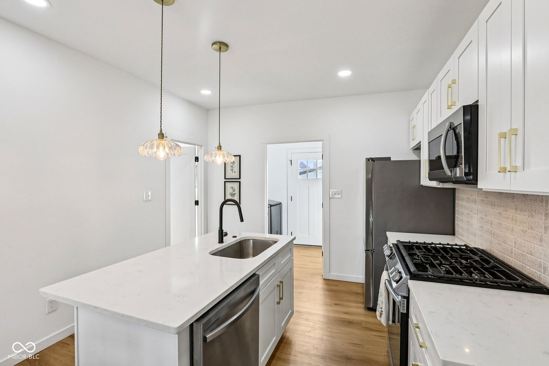 Bright kitchen featuring a central island with a stainless steel sink and matte black faucet, pendant lighting, white cabinetry with gold-tone hardware, stainless steel appliances, and wood-finish flooring