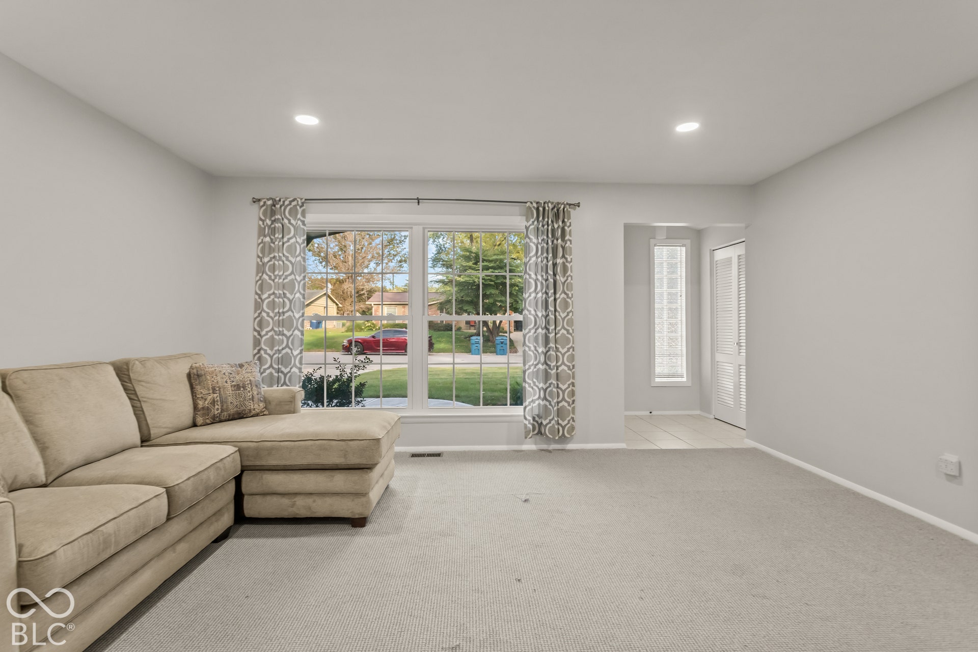 Recessed ceiling lighting, light gray wall paint, large window with white trim, gray carpet, and a tiled entry area