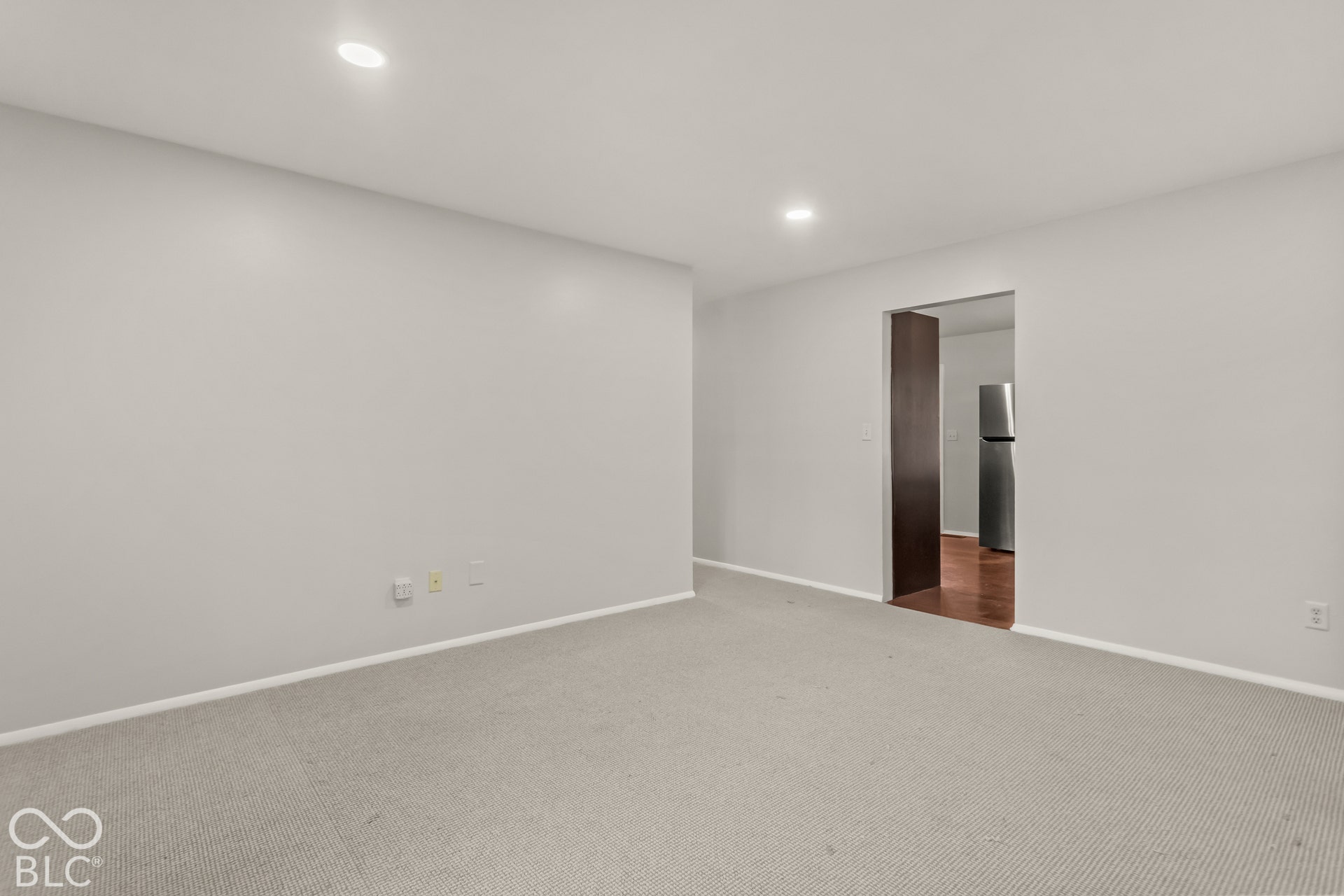 Recessed ceiling lighting, light gray wall paint, light-toned carpeting, wood-finish doorway trim, and a stainless steel refrigerator