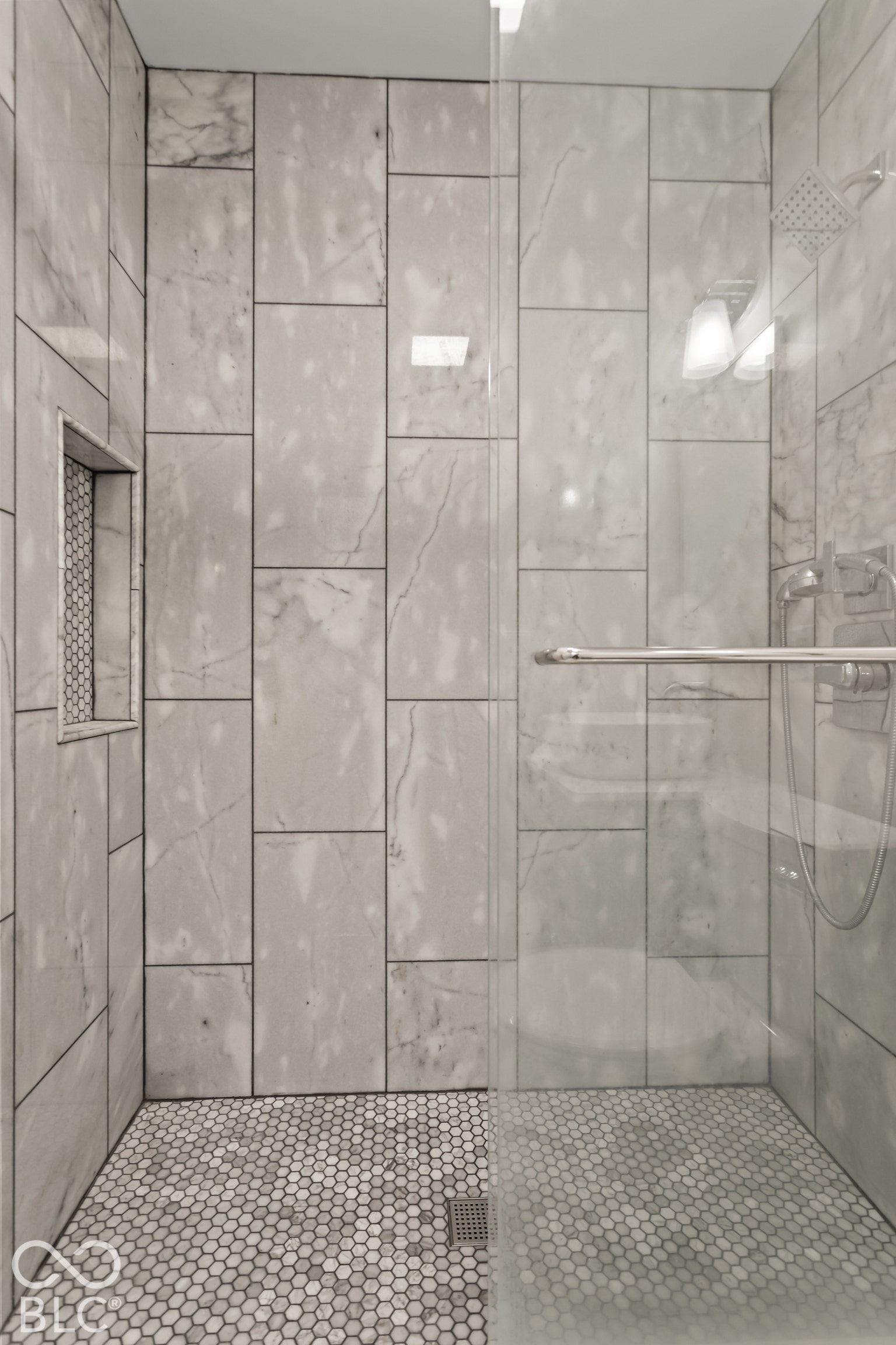 Shower featuring large format wall tiling, a built-in shower niche, a frameless glass enclosure, a rainfall shower head, and hexagonal floor tiling
