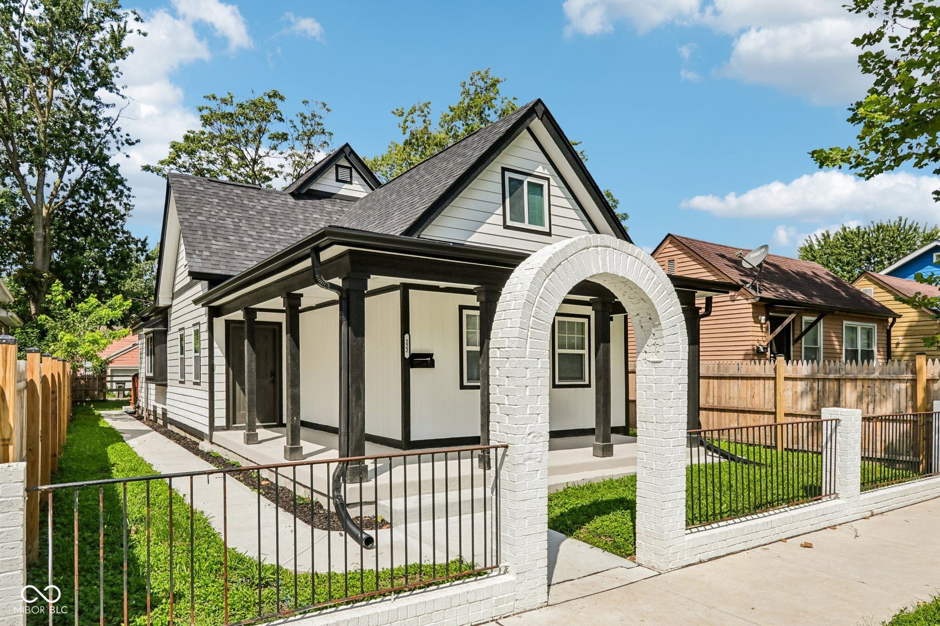 White siding exterior with dark trim accents, covered front porch, brick archway entry, and black metal fencing