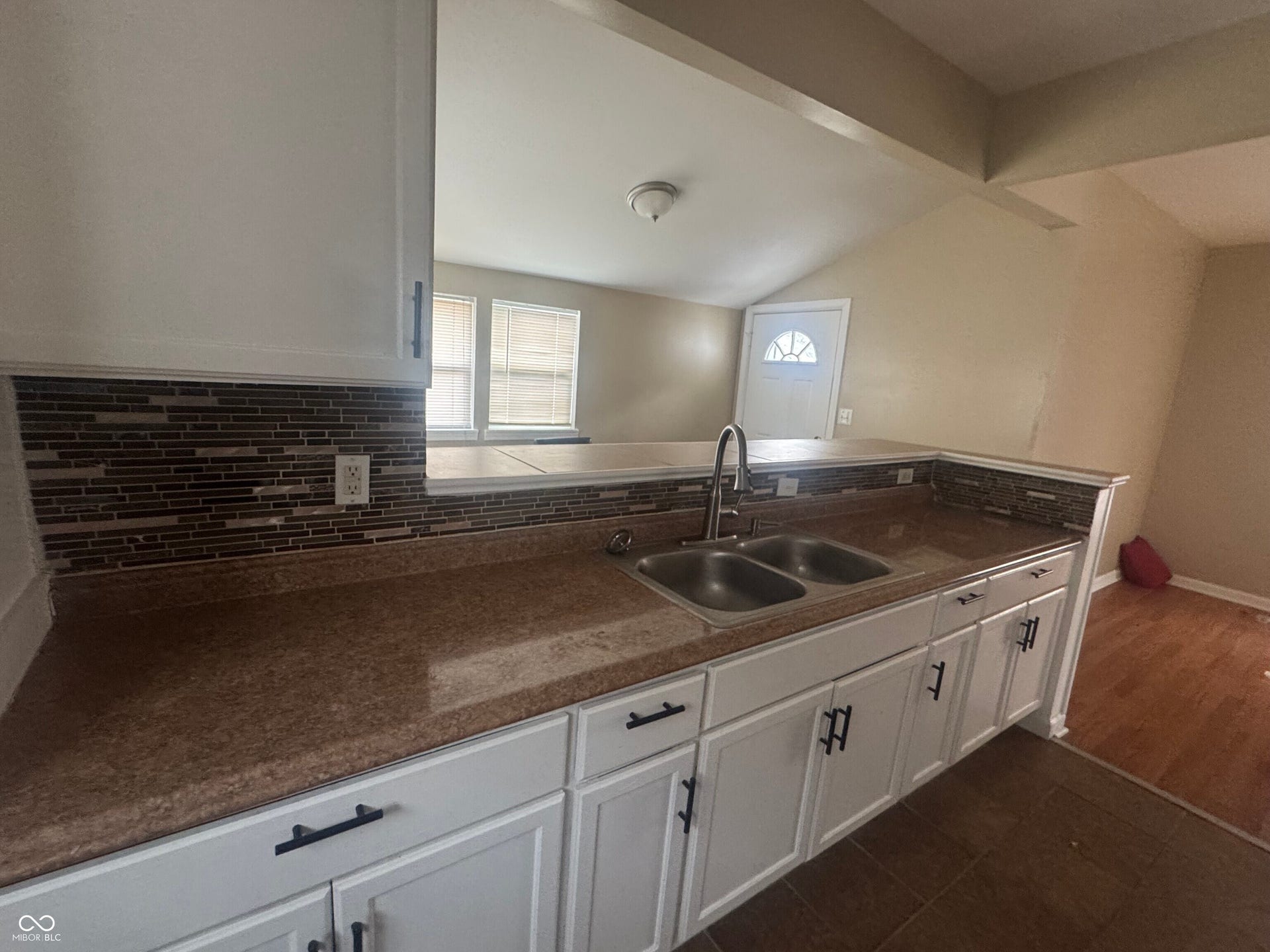 Virtually Altered - Modern kitchen featuring white cabinetry with black hardware, granite-style countertops, a double-basin stainless steel sink with contemporary faucet, and a linear glass tile backsplash