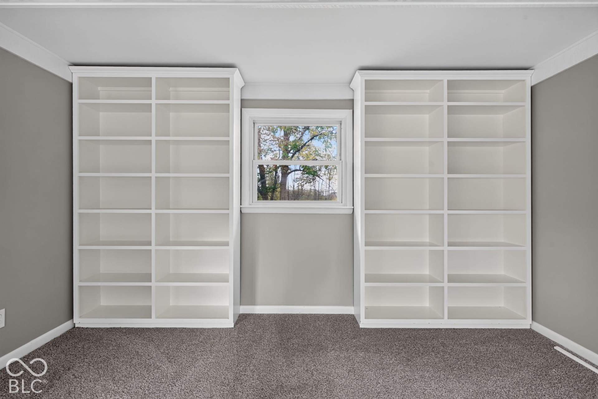 Floor-to-ceiling built-in white shelving, crown molding, and a central window with exterior tree-lined scenery