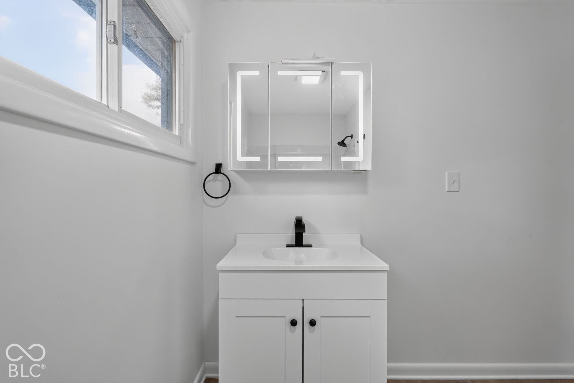 White vanity with a rectangular sink, matte black faucet, integrated lighting medicine cabinet, and wall-mounted towel ring
