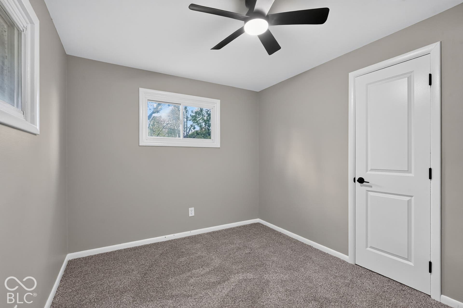 Neutral wall finish, gray carpeting, sliding window with white trim, five-blade ceiling fan with integrated lighting, and white panel door with matte black hardware