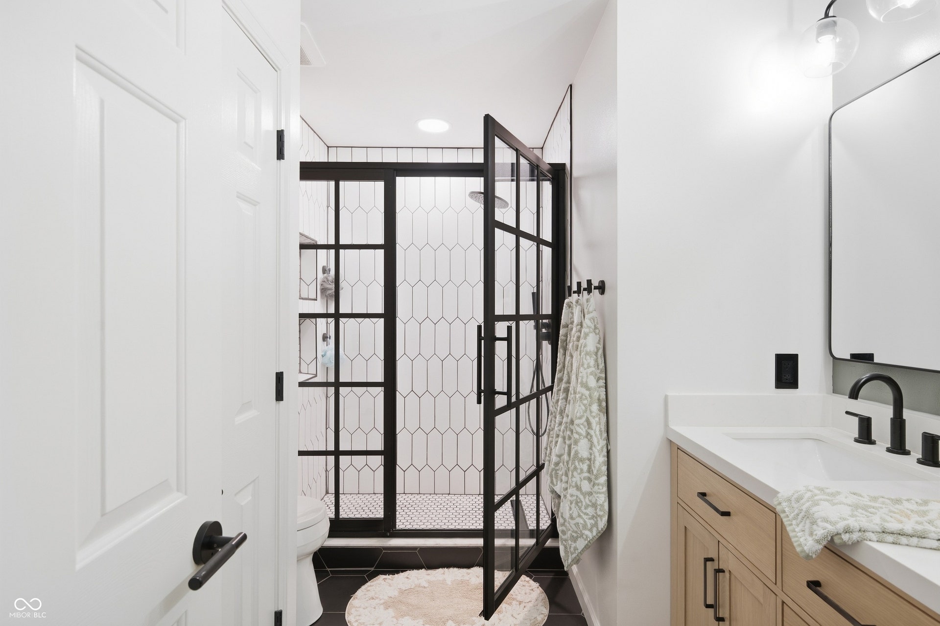 Shower with black grid glass enclosure and hexagonal tile, wood-finish vanity with countertop, and black hardware