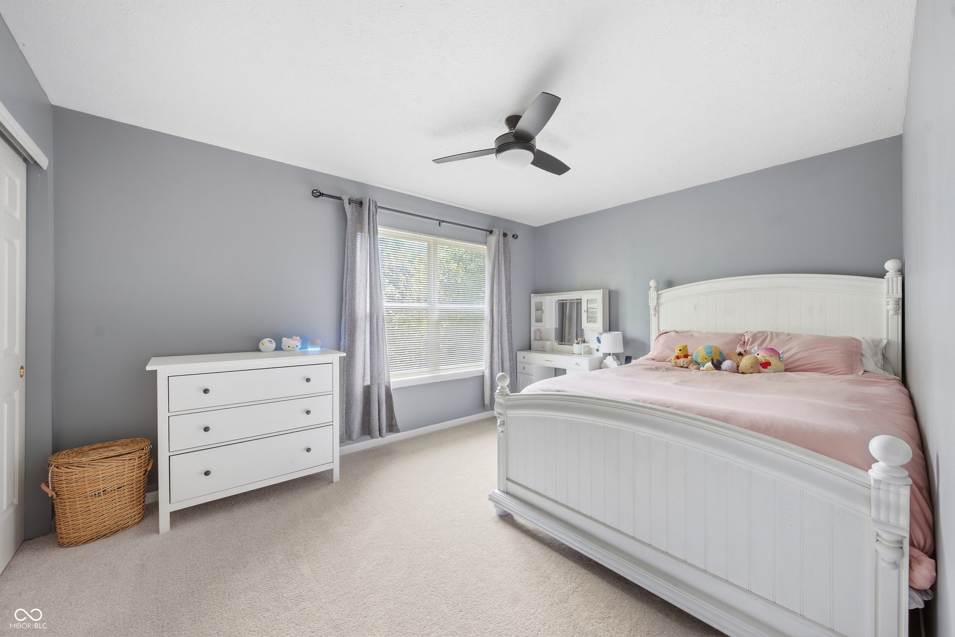 Light gray wall paint, textured ceiling, light-colored carpet flooring, a window with a dark rod and light curtains, and a ceiling fan with integrated lighting