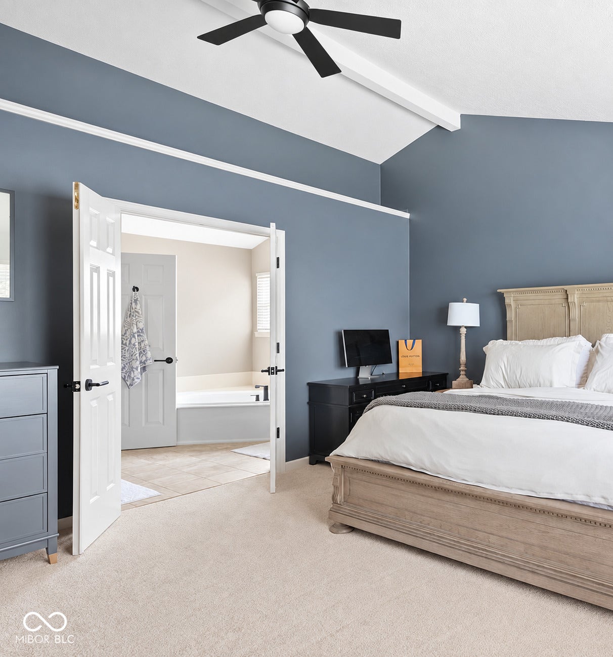 Vaulted ceiling with exposed beam, modern ceiling fan, French doors, plush carpeting, and an en-suite bathroom featuring a built-in tub