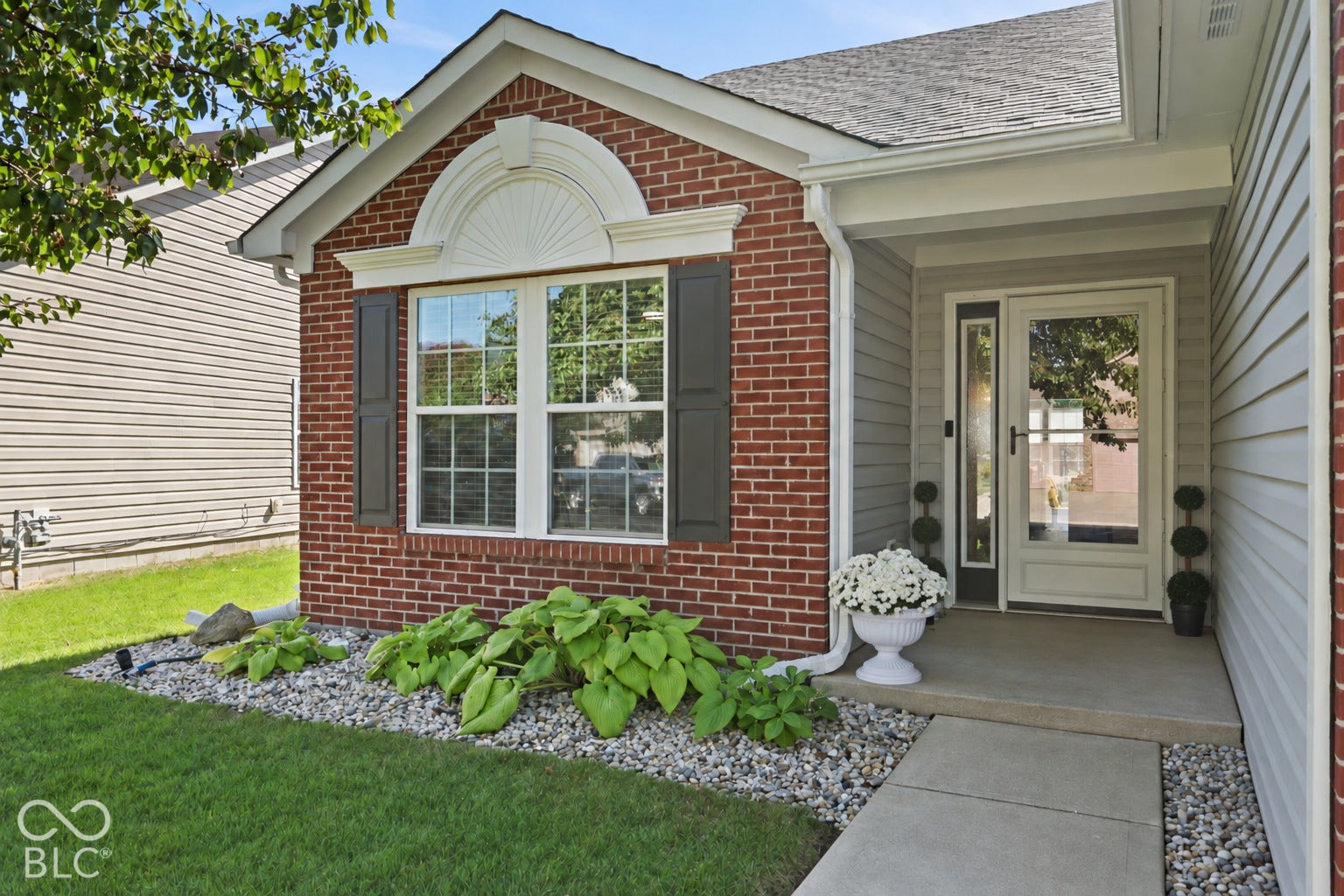 Brick facade exterior featuring a decorative arched window header, grey shutters, a concrete walkway, and gravel landscaping