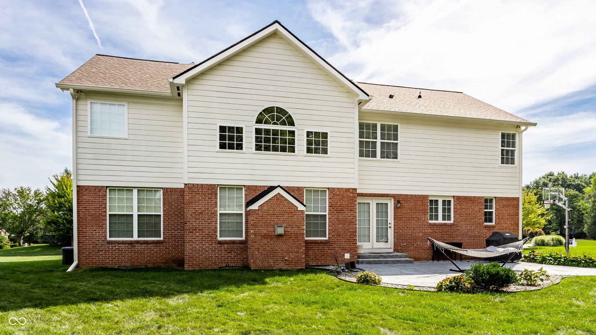 Two-story exterior with brick and siding, featuring multiple windows, a second-story arched window, and a ground-level patio with double doors