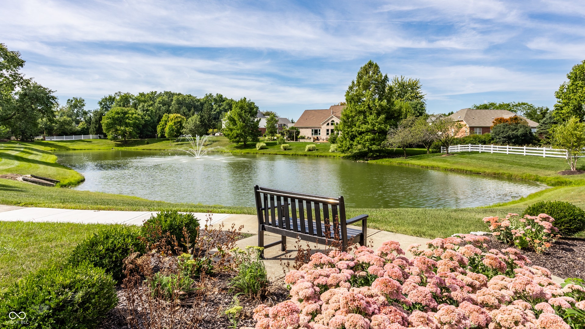Pond with a fountain, surrounded by green landscaping, mature trees, and a white picket fence