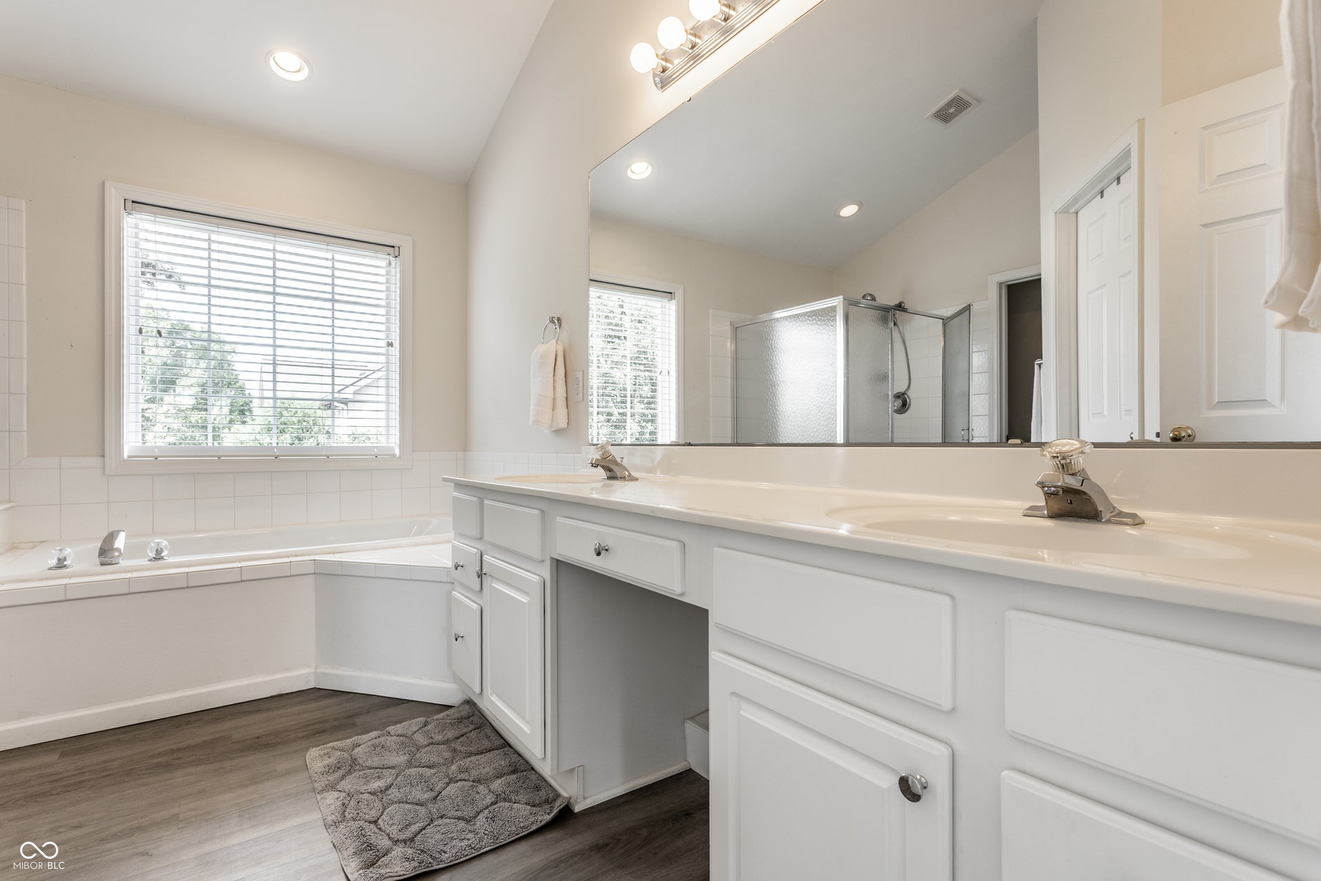 Dual vanity with white cabinetry and integrated sinks, a corner soaking tub with white tile surround, a glass-enclosed shower.