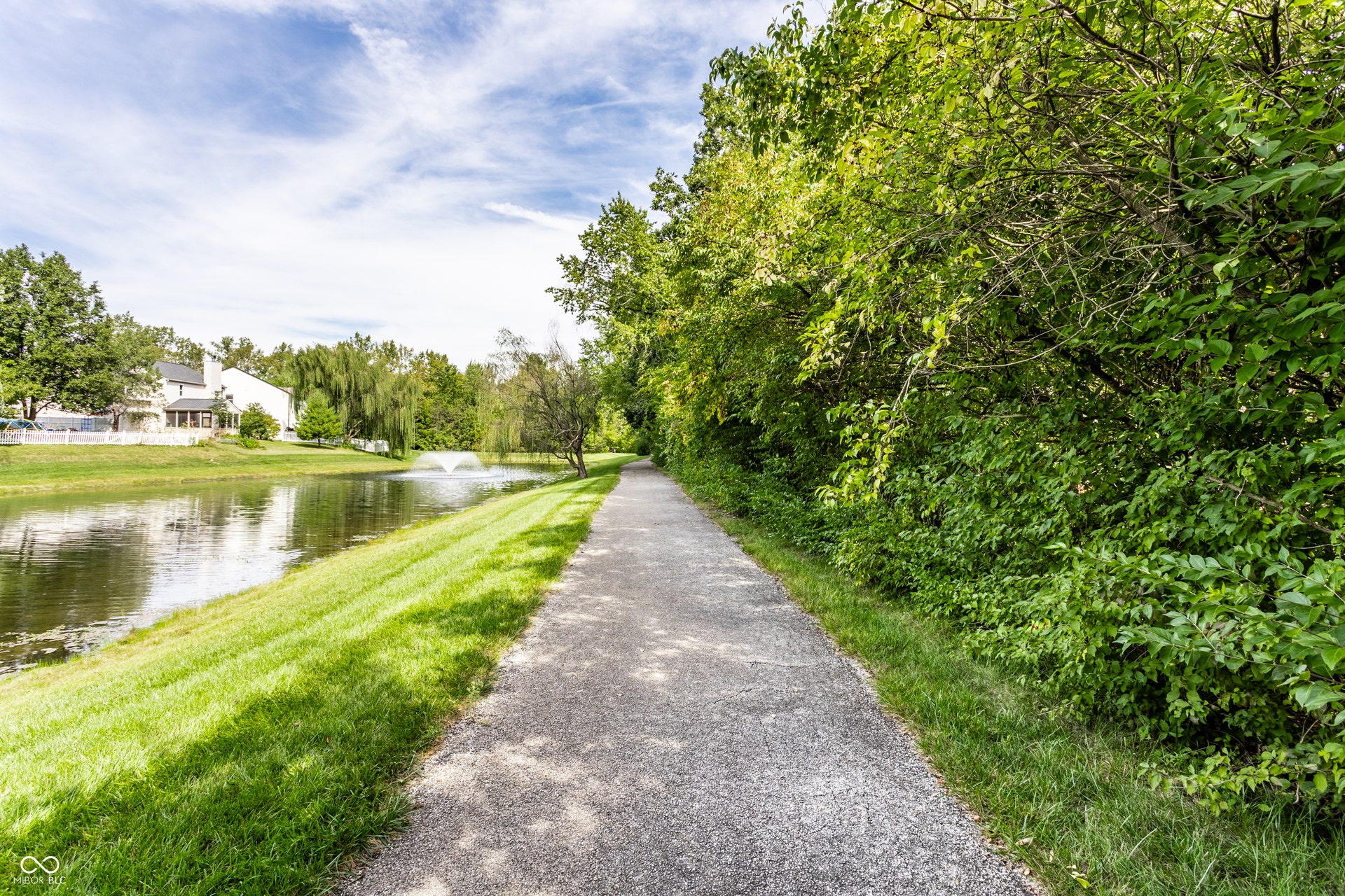 Paved pathway into the nature preserve, bordered by manicured lawn and mature trees, featuring a pond with a fountain