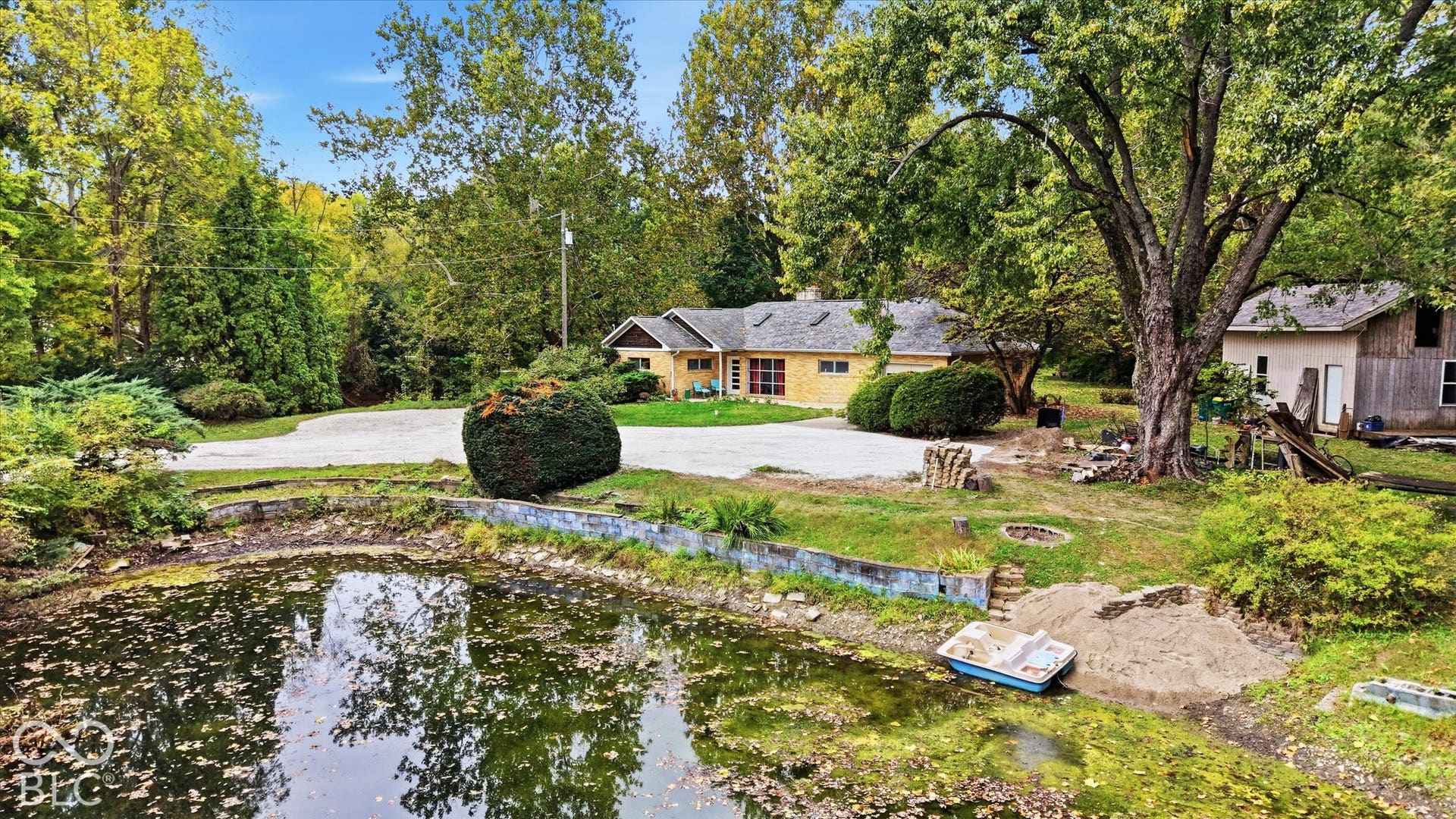 Stone exterior residence with a gravel driveway, mature trees, and a pond