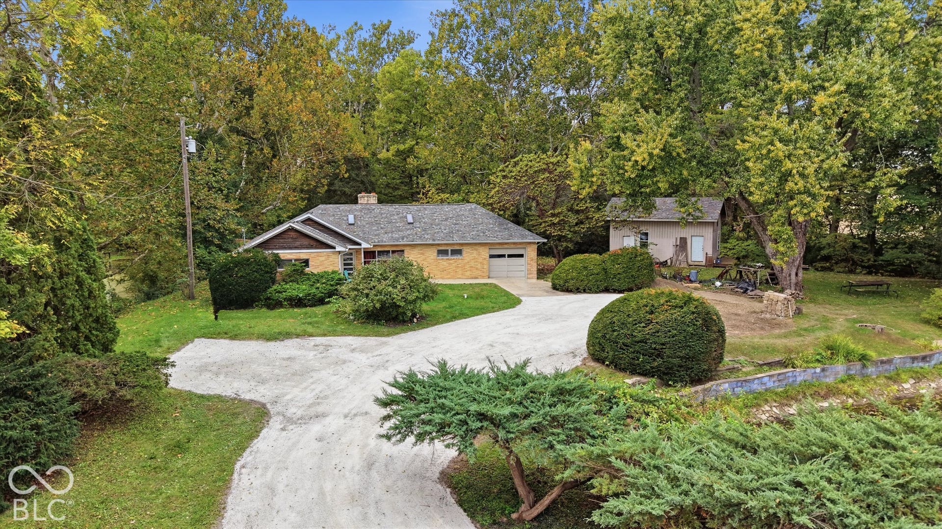 Single-story brick exterior with an attached garage, featuring a gravel driveway, mature trees, and an additional detached structure