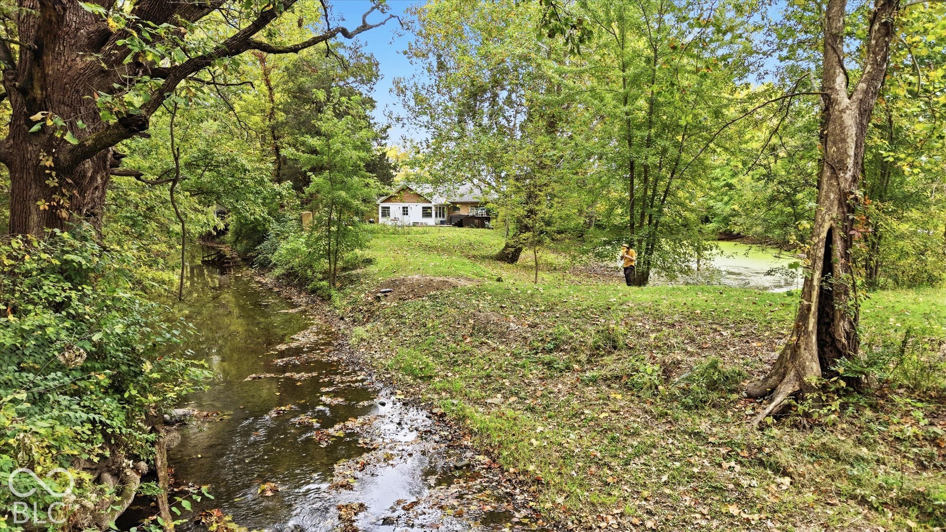 White exterior structure situated on a grassy incline, bordered by mature trees and a natural stream