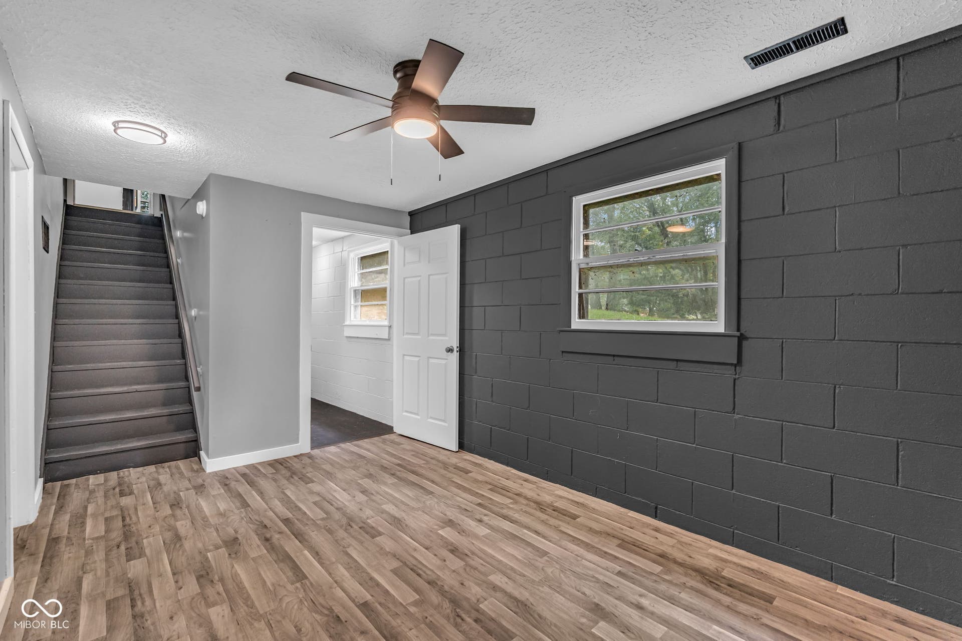Wood-finish flooring, staircase with dark treads and handrail, ceiling fan with integrated lighting, window with horizontal blinds, and a white interior door