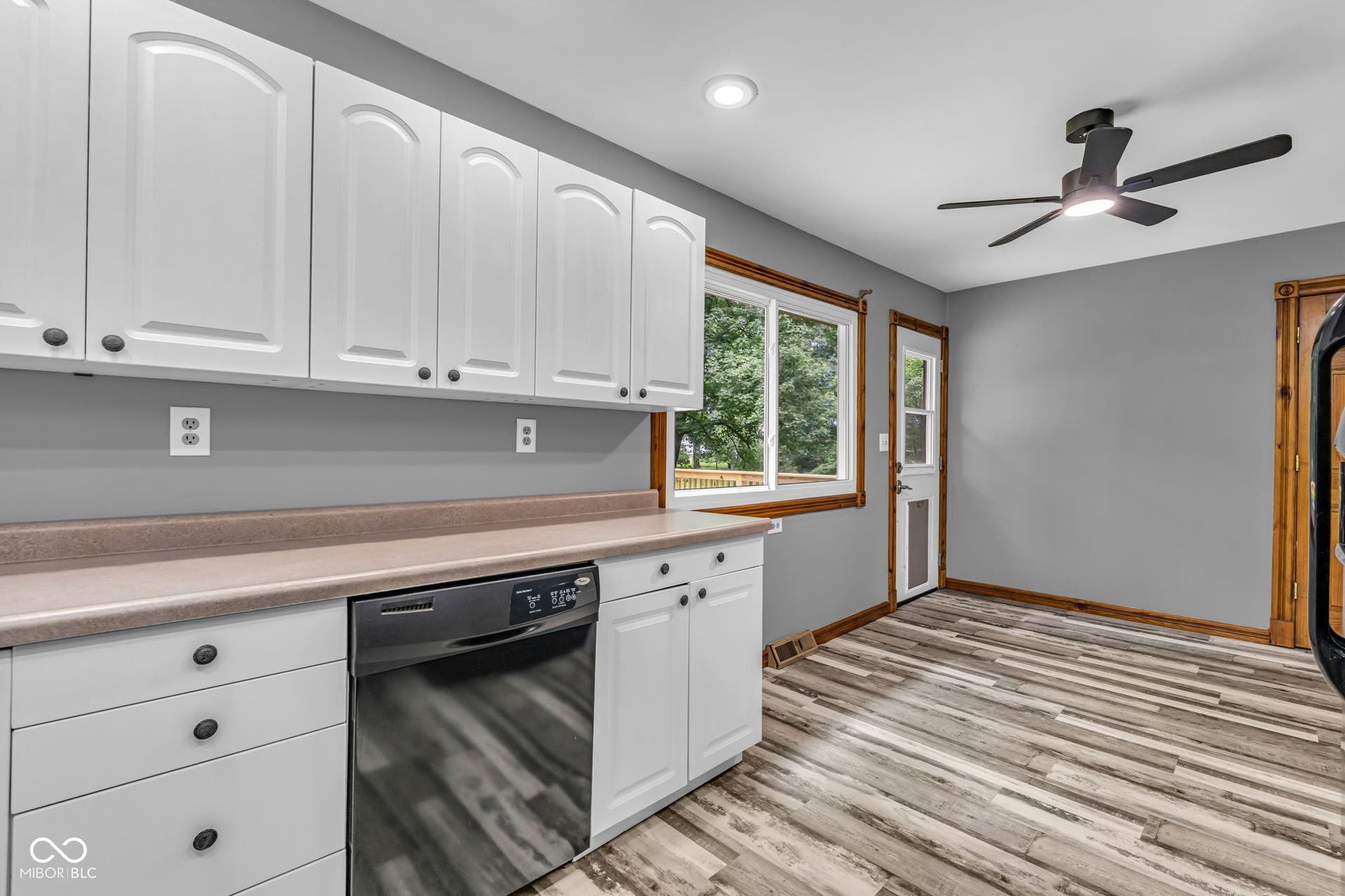 White upper and lower cabinetry, stainless steel dishwasher, light-toned countertop, wood-finish flooring, and a ceiling fan with integrated lighting
