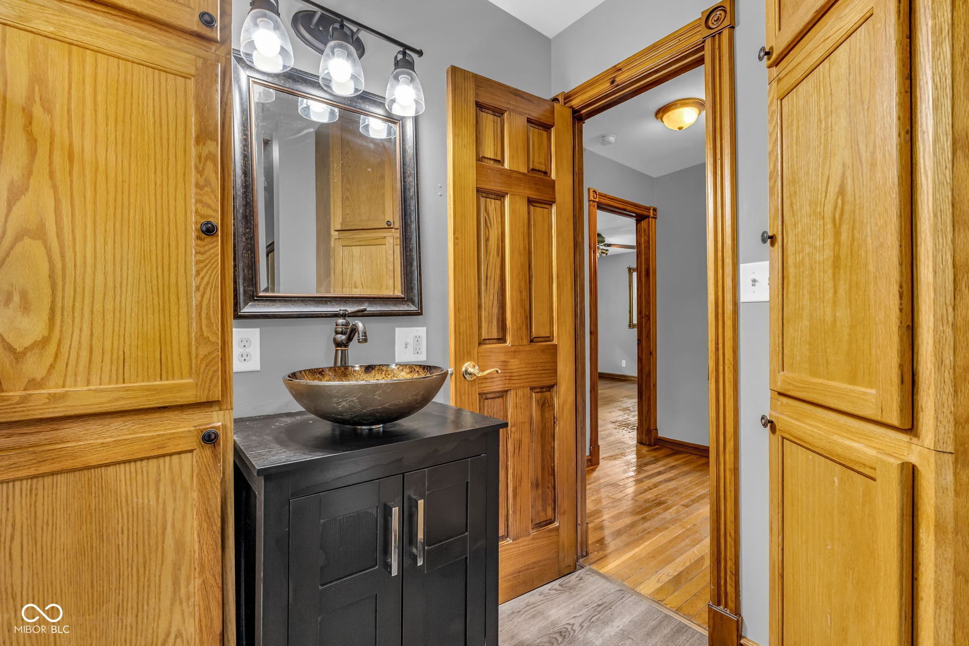 Wood-finish flooring with a vessel sink, dark wood vanity, framed mirror, and integrated lighting