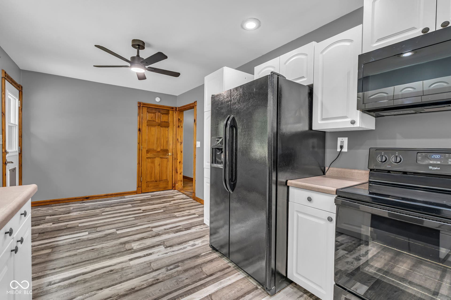 Kitchen area featuring wood-finish flooring, white cabinetry, integrated black appliances, light-toned countertops, and a ceiling fan with integrated lighting