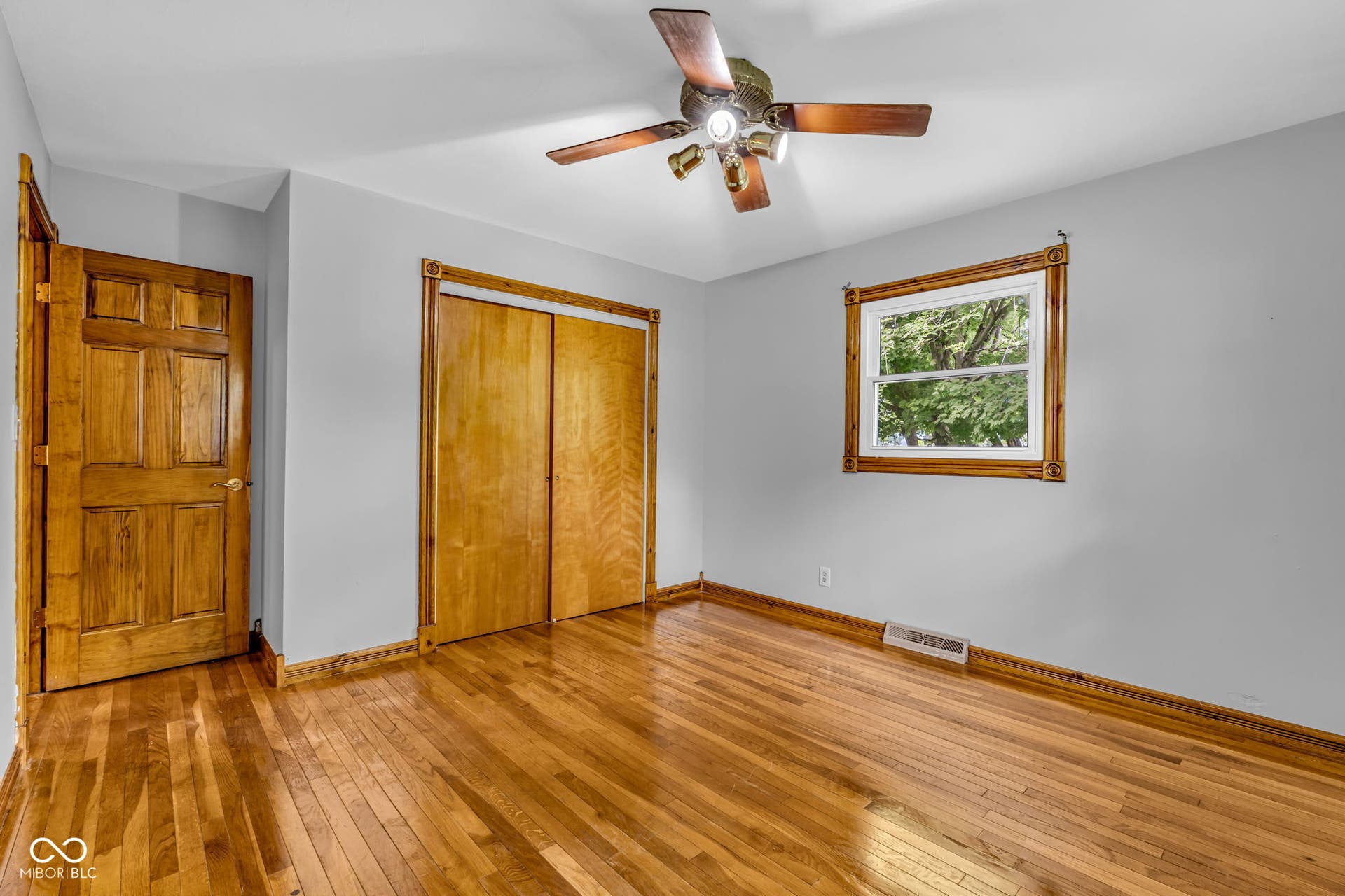 Hardwood flooring throughout, a ceiling fan with integrated lighting, a paneled wooden door, a double-door closet with wooden bi-fold doors, and a window with wooden trim