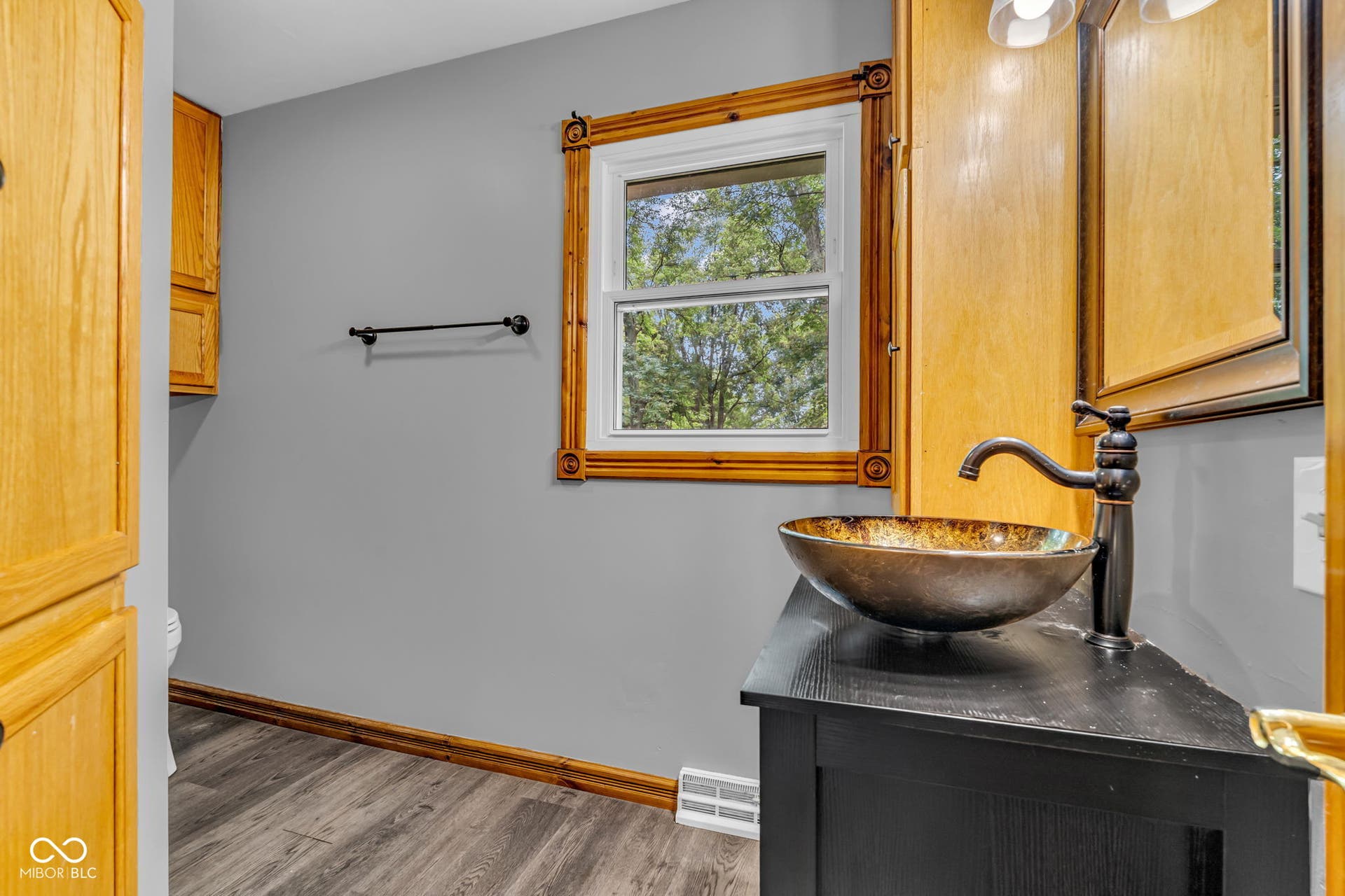 Wood-finish flooring with coordinating wood trim, custom wood window framing, vessel sink with an oil-rubbed bronze faucet, and a wall-mounted light fixture