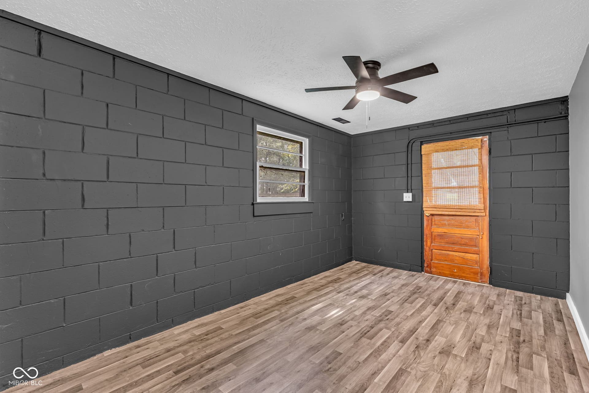 Wood-finish flooring, dark-painted block walls, multi-pane window, ceiling fan with integrated lighting, and a unique wood-paneled door with an upper screen panel
