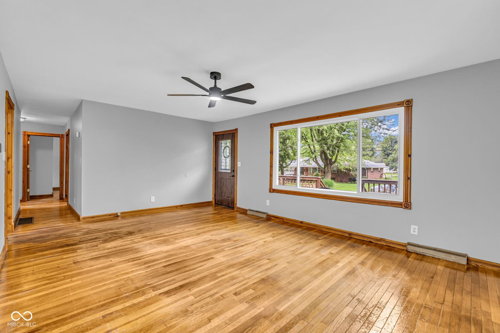 Hardwood flooring throughout, large picture window with wood trim, ceiling fan with light, and a solid wood entry door with glass insert