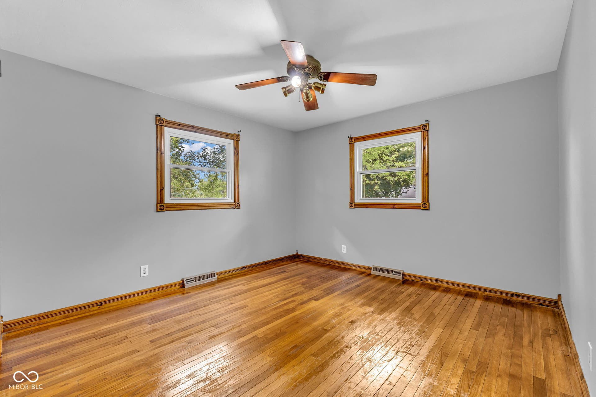 Hardwood flooring, two windows with decorative wood trim, ceiling fan, and light gray wall finish