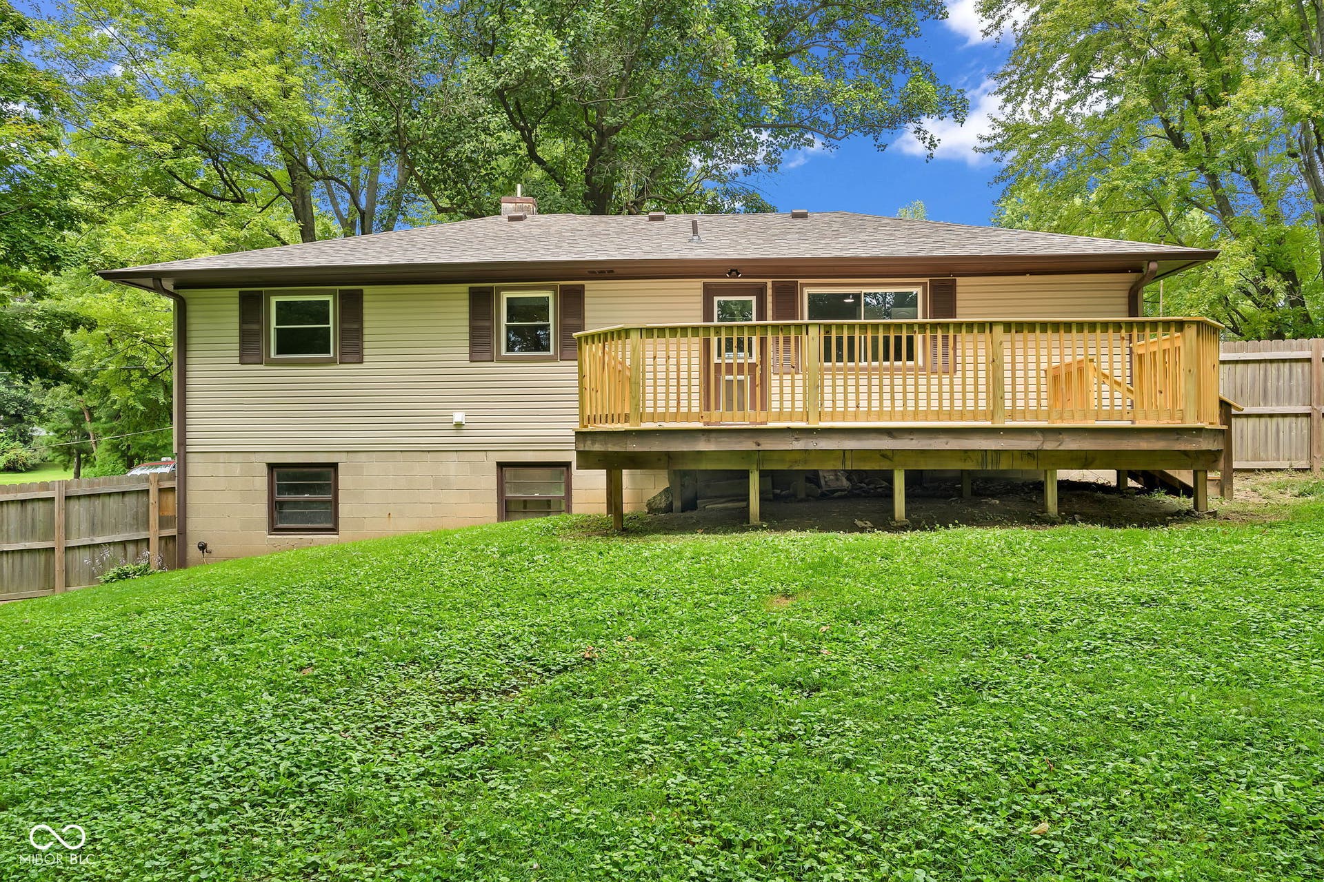 Expansive rear deck, tan horizontal siding, brown window shutters, fenced perimeter, and a sloped lawn