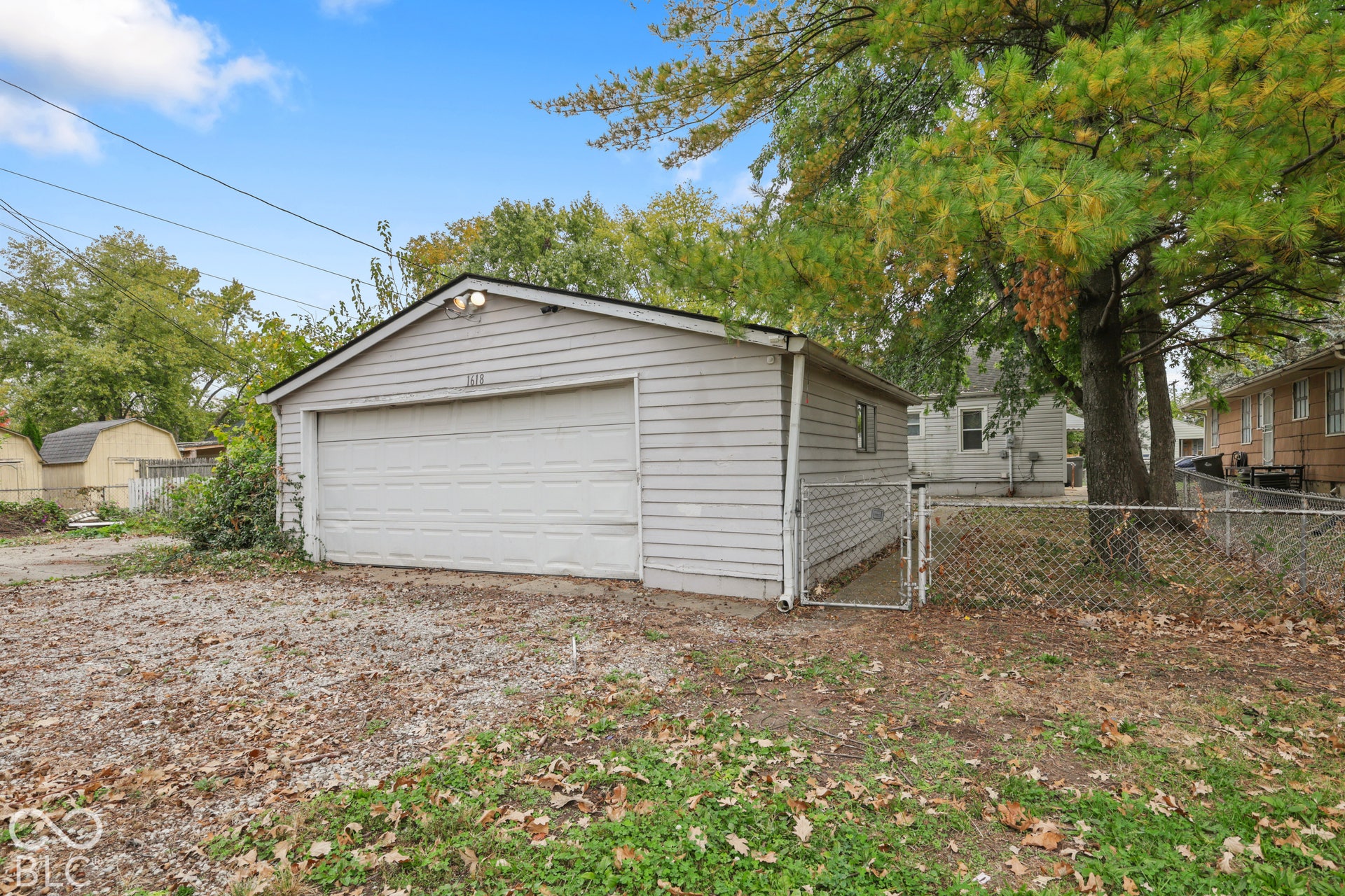 Detached two-car garage featuring horizontal siding, a roll-up sectional door, exterior mounted lighting, and chain-link perimeter fencing