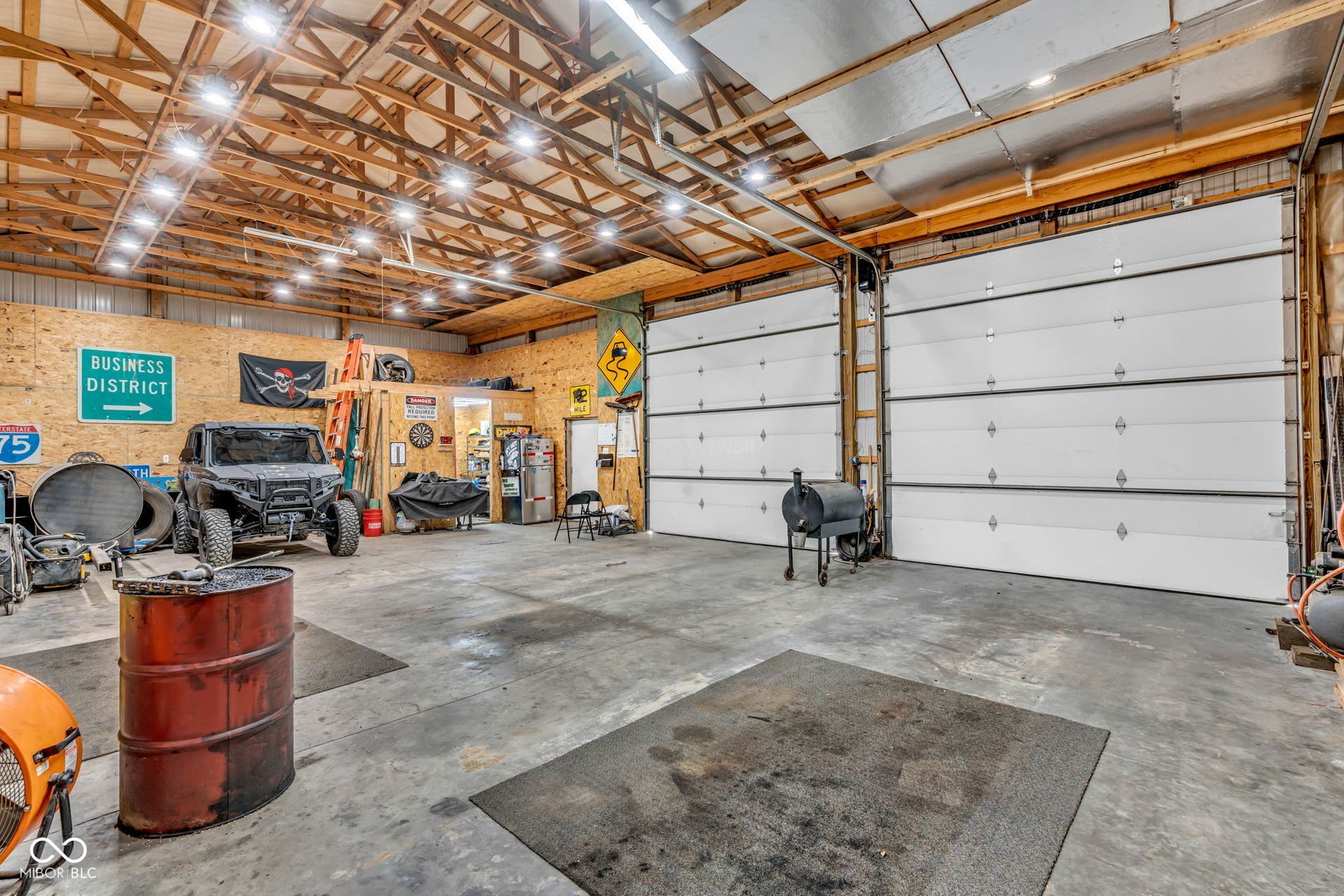 5432 W Whittier Drive, Knightstown, IN 46148 Concrete flooring, exposed wood beam ceiling, multiple overhead light fixtures, two large roll-up doors, and plywood wall finishes
