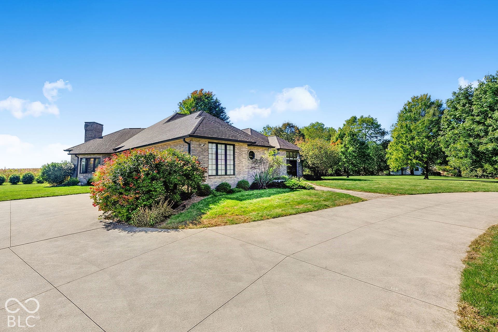Stone facade residence featuring a dark shingle roof, chimney, large concrete driveway, and mature landscaping