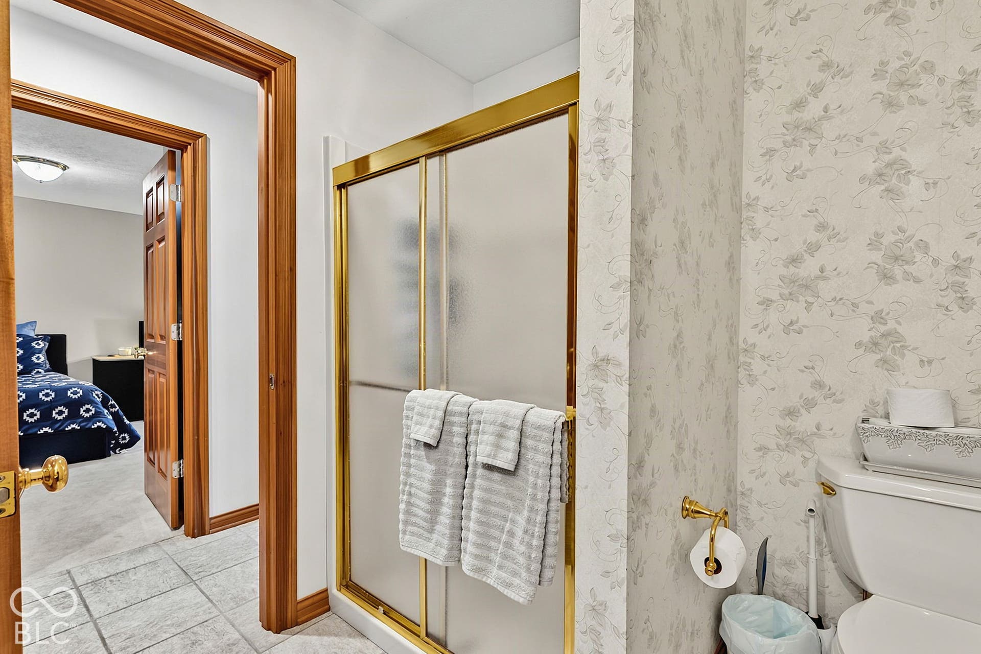 Enclosed shower with gold-tone frame, tiled flooring, wood-finish door frames, and textured wallpaper