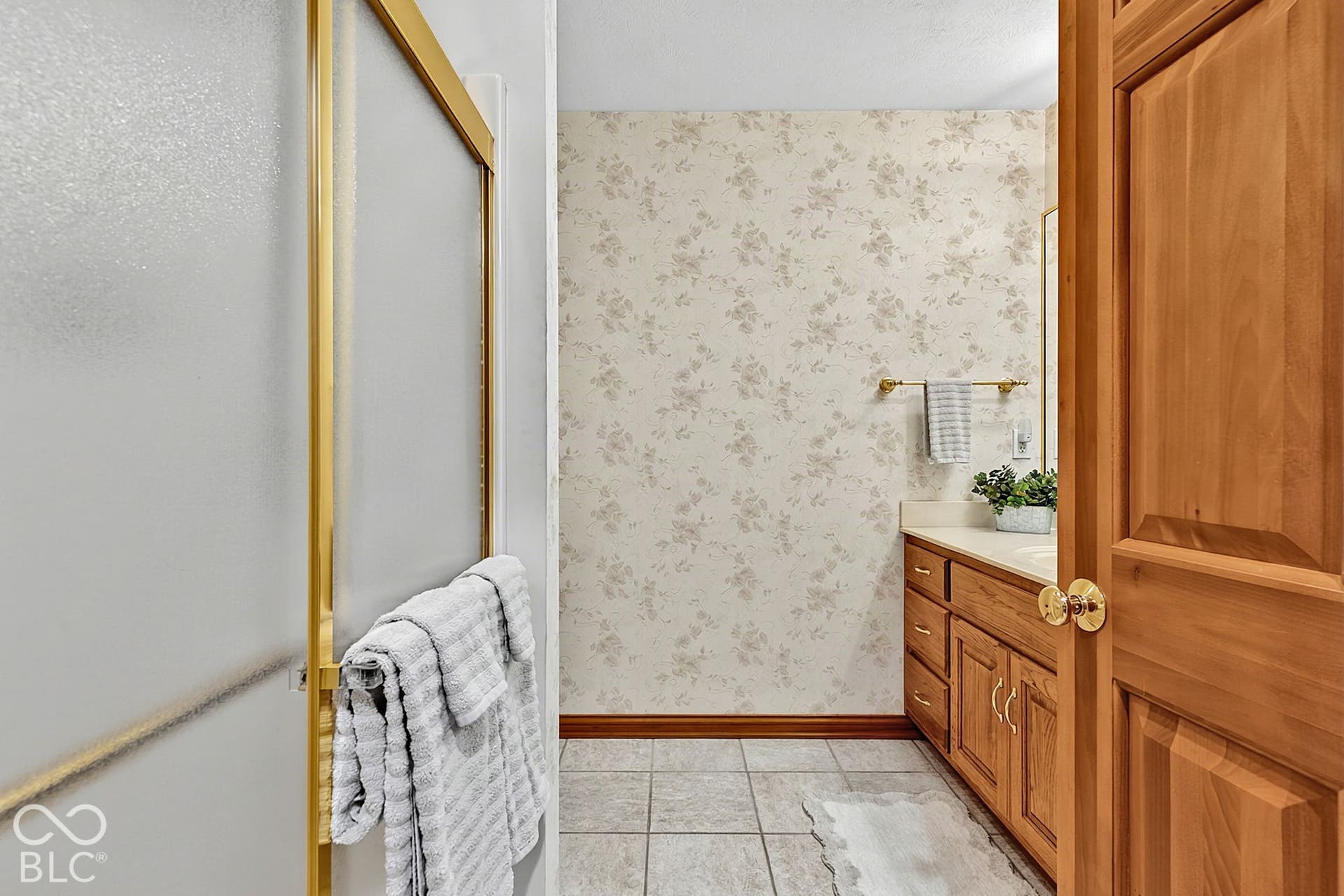 Frosted glass shower enclosure with gold-tone framing, wood-finish vanity with a light-toned countertop, and patterned wallpaper