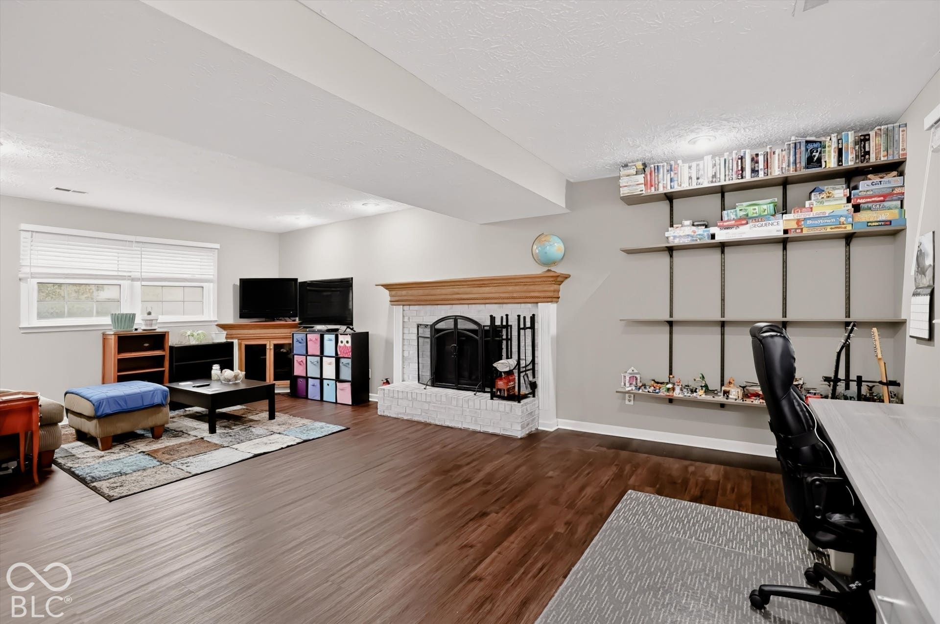 Living area featuring wood-finish flooring, white-painted brick fireplace with a wood mantel, and wall-mounted shelving units