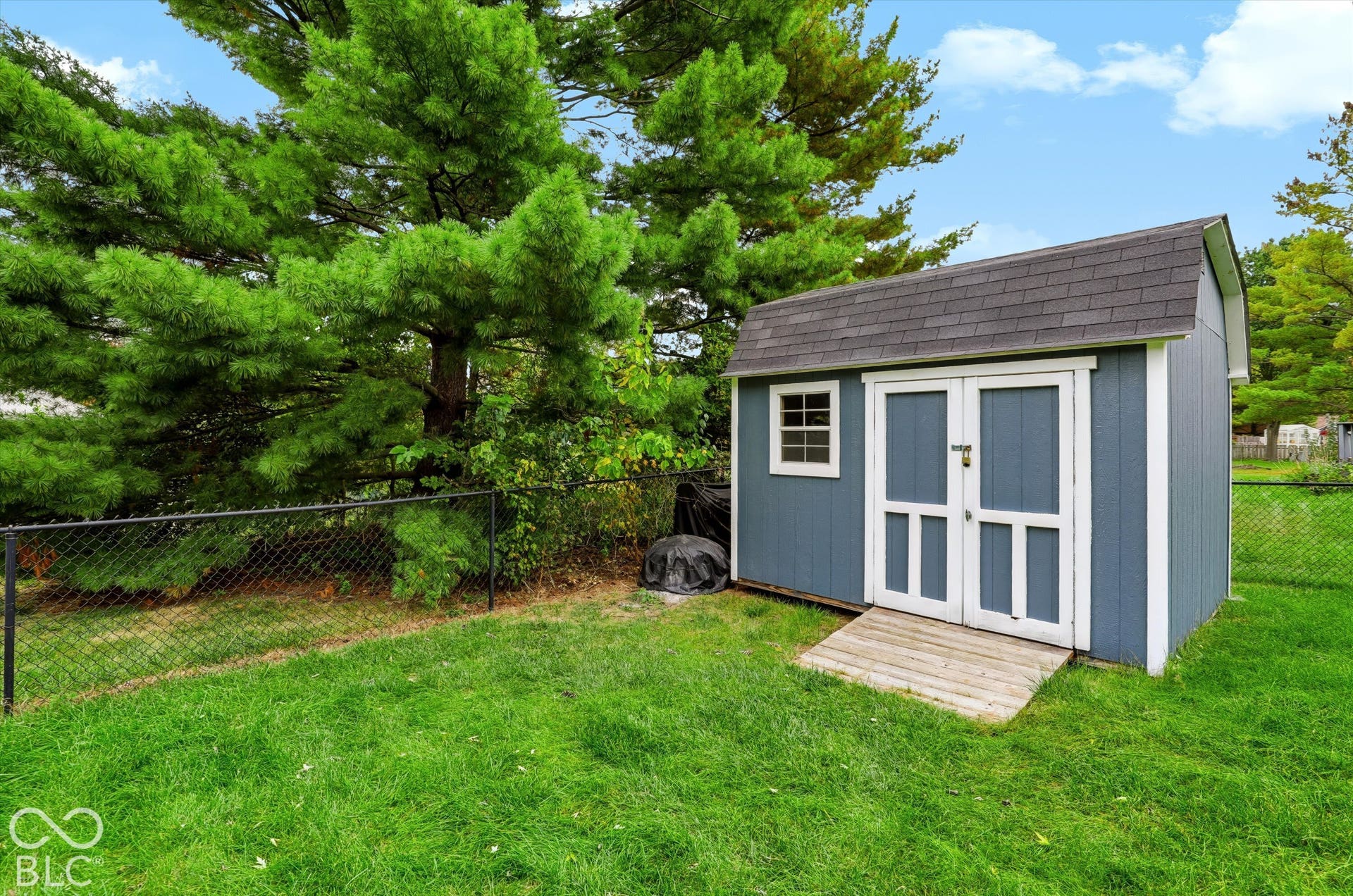 Backyard storage shed featuring a gambrel roof, double-door entry, wood ramp, and integrated window, surrounded by chain-link fencing and mature evergreen trees