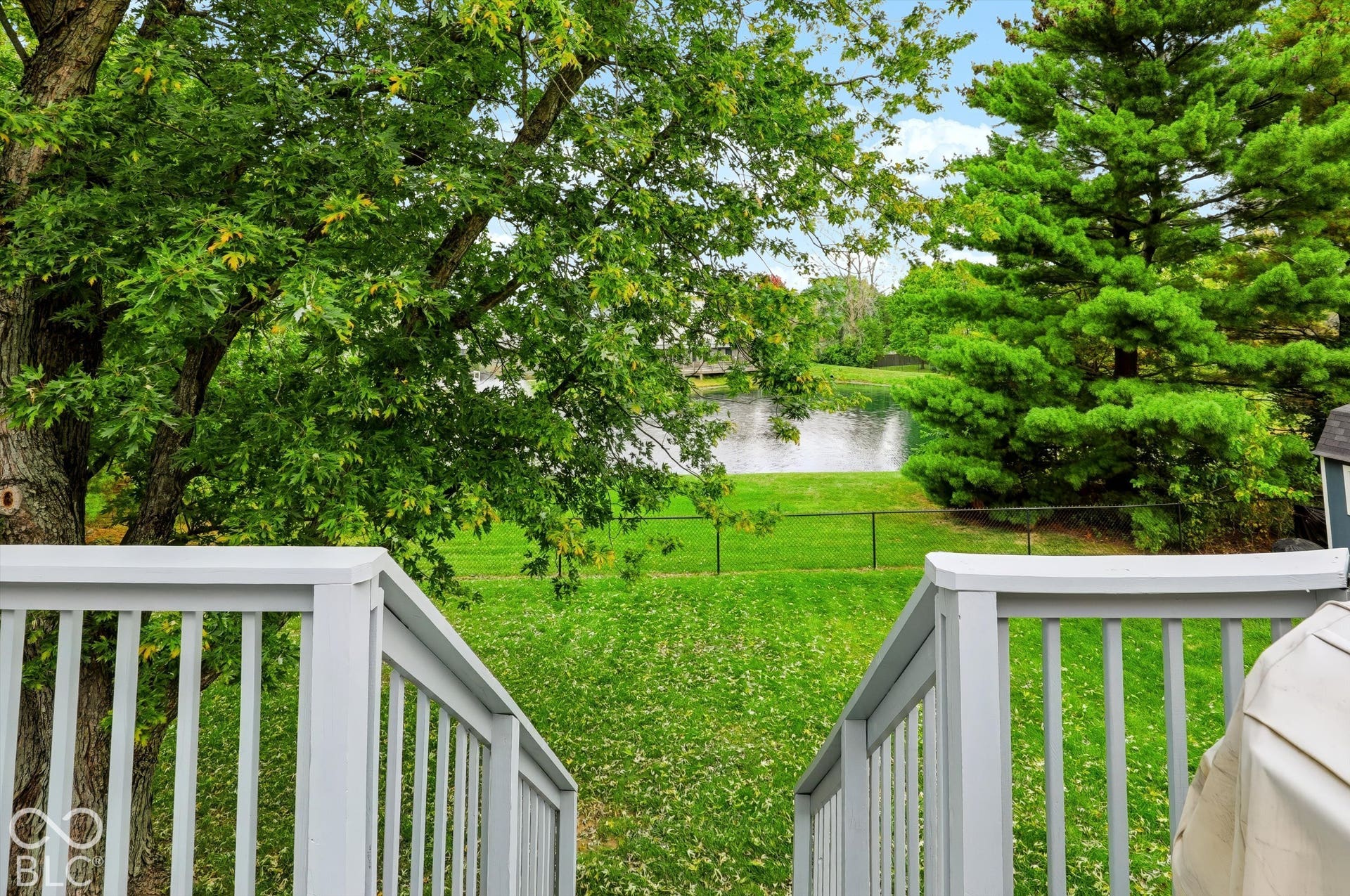 Elevated deck featuring painted railings and staircases overlooking a manicured lawn, mature trees, and a pond