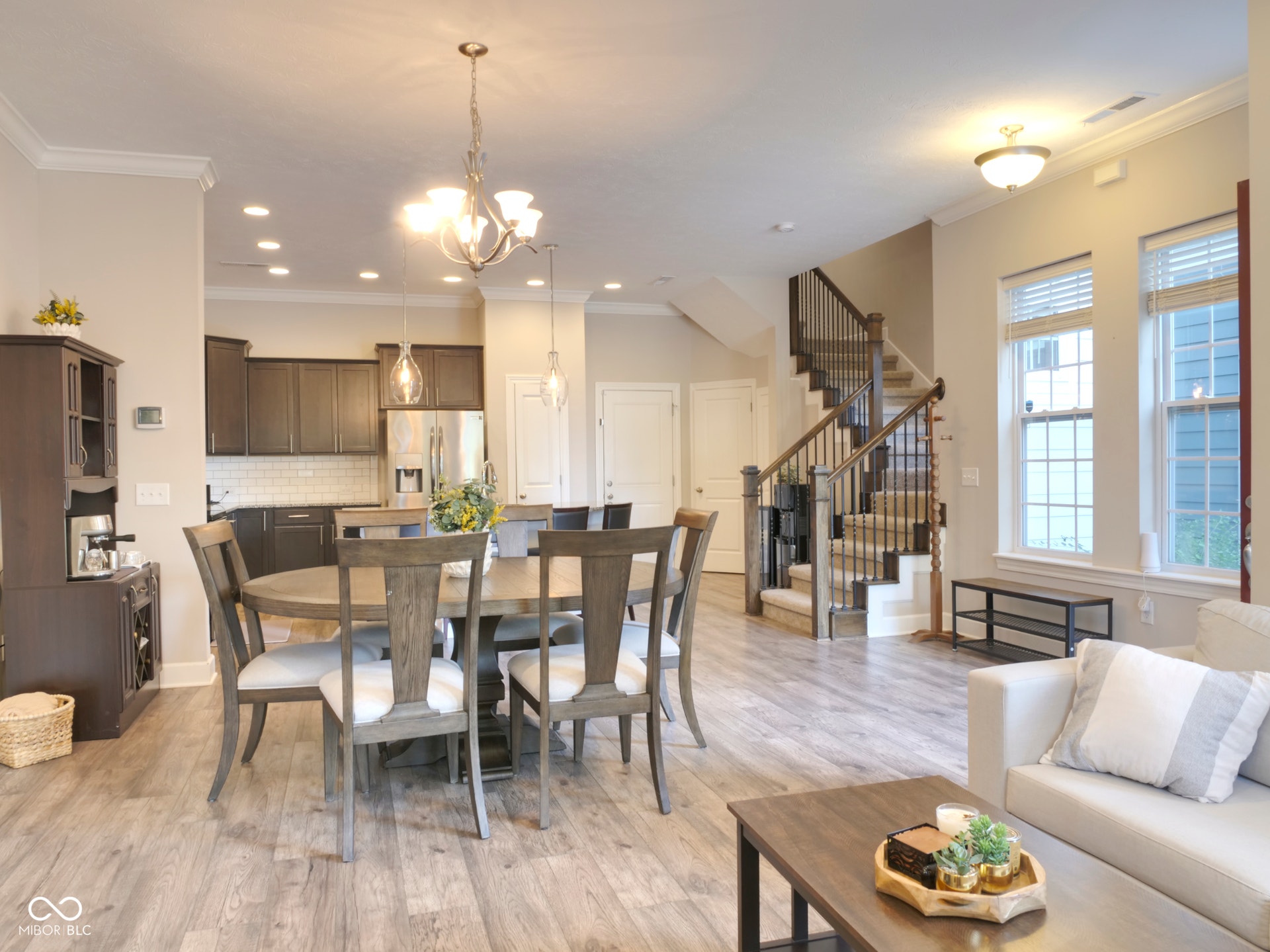 Wood-finish flooring throughout, kitchen with dark cabinetry and stainless steel appliances, open-concept living space, staircase with dark wood banister and carpeted treads, multiple windows providing natural light