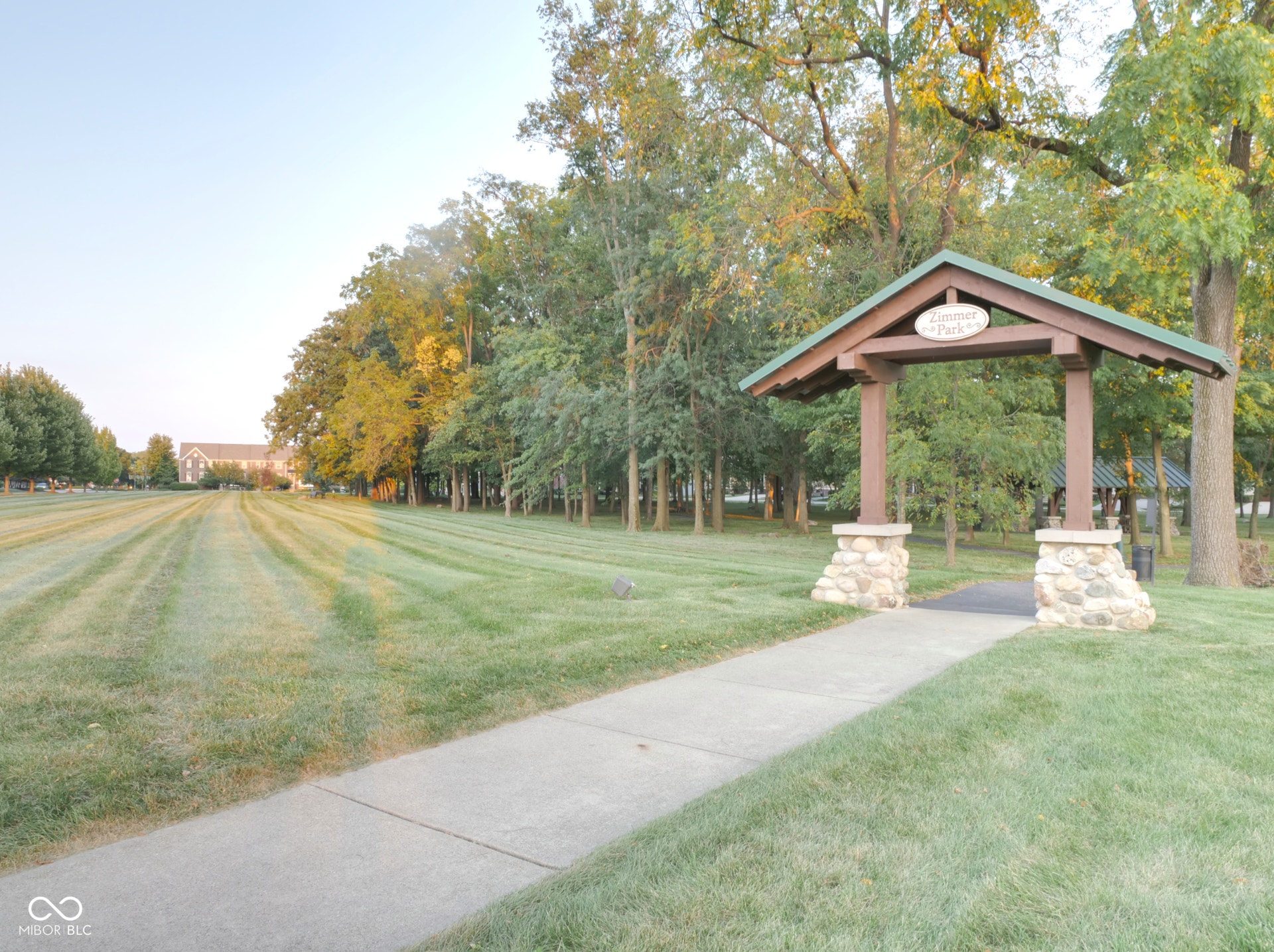 Expansive green space featuring a paved walkway, mature trees, and a decorative wooden archway with stone bases
