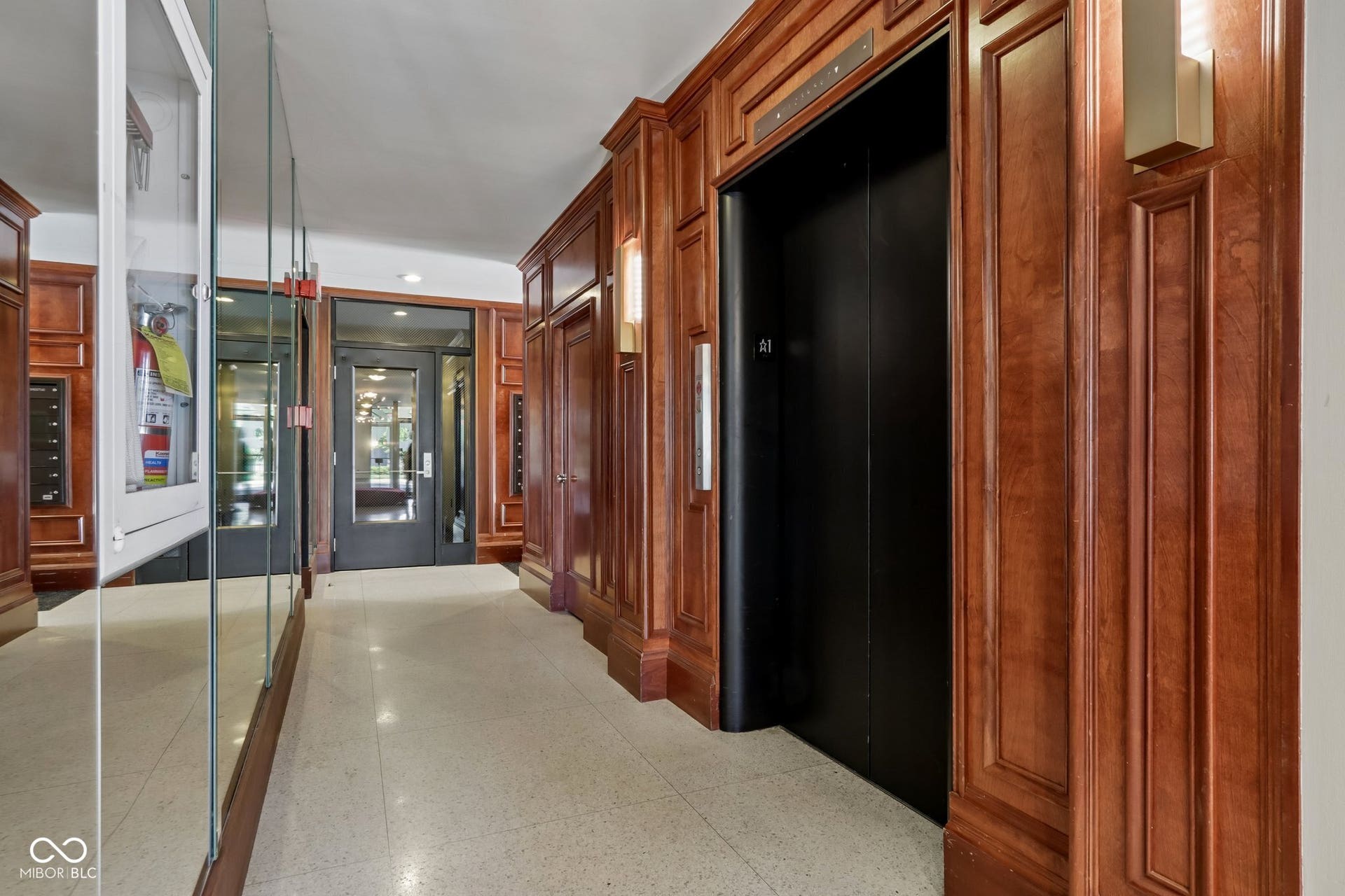 Hallway featuring rich wood paneling, an elevator with a black door, wall-mounted light fixtures, and terrazzo flooring