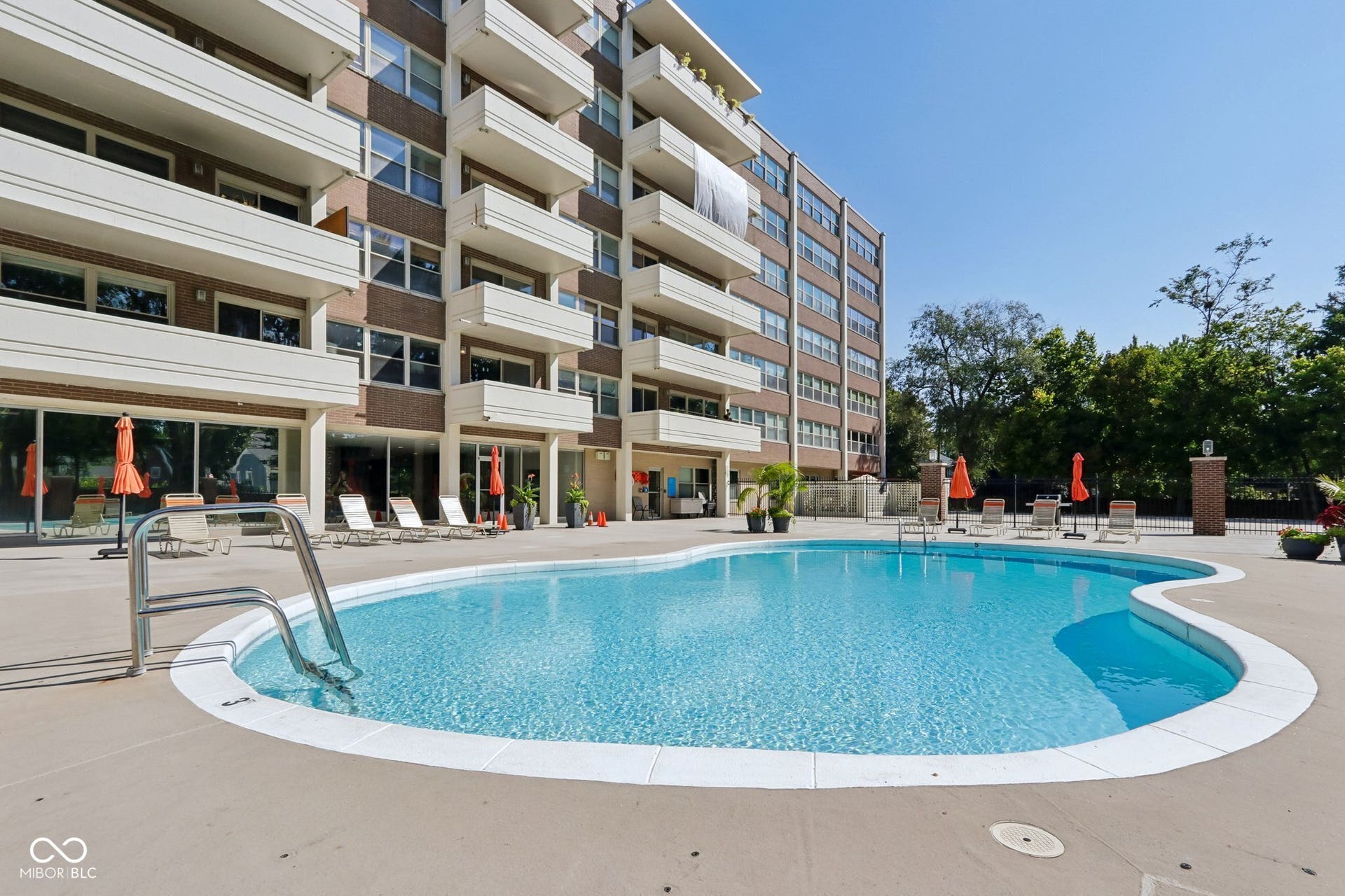 Curved outdoor swimming pool with a concrete surround, featuring a multi-story building with private balconies and a brick facade