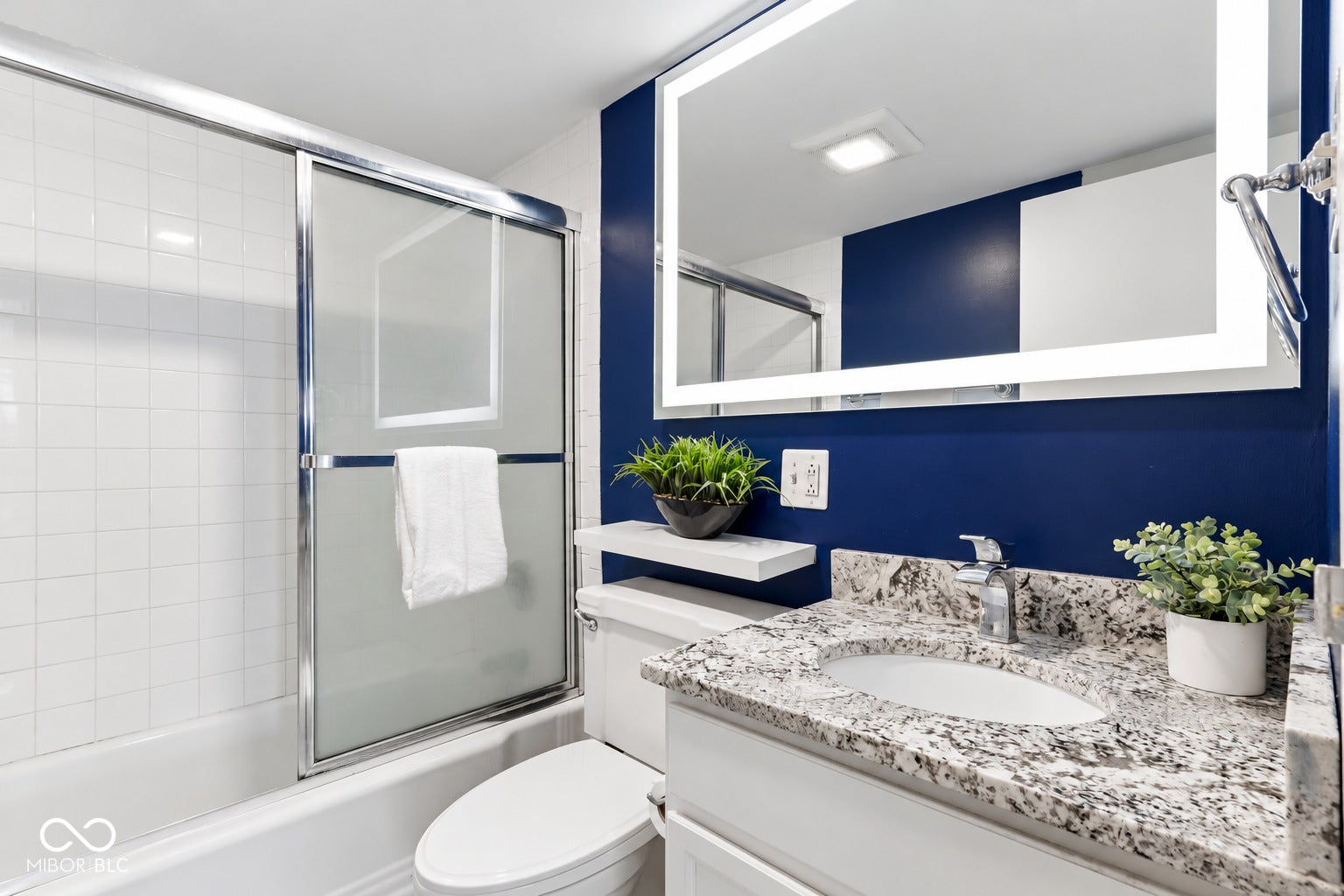 Tub with sliding glass shower doors, white subway tile surround, vanity with granite-finish countertop, single basin sink, and an illuminated mirror