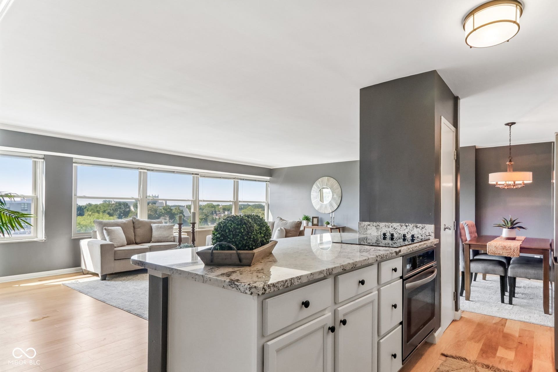 Open-concept living area with large windows, wood-finish flooring, and a kitchen island featuring a built-in cooktop and oven