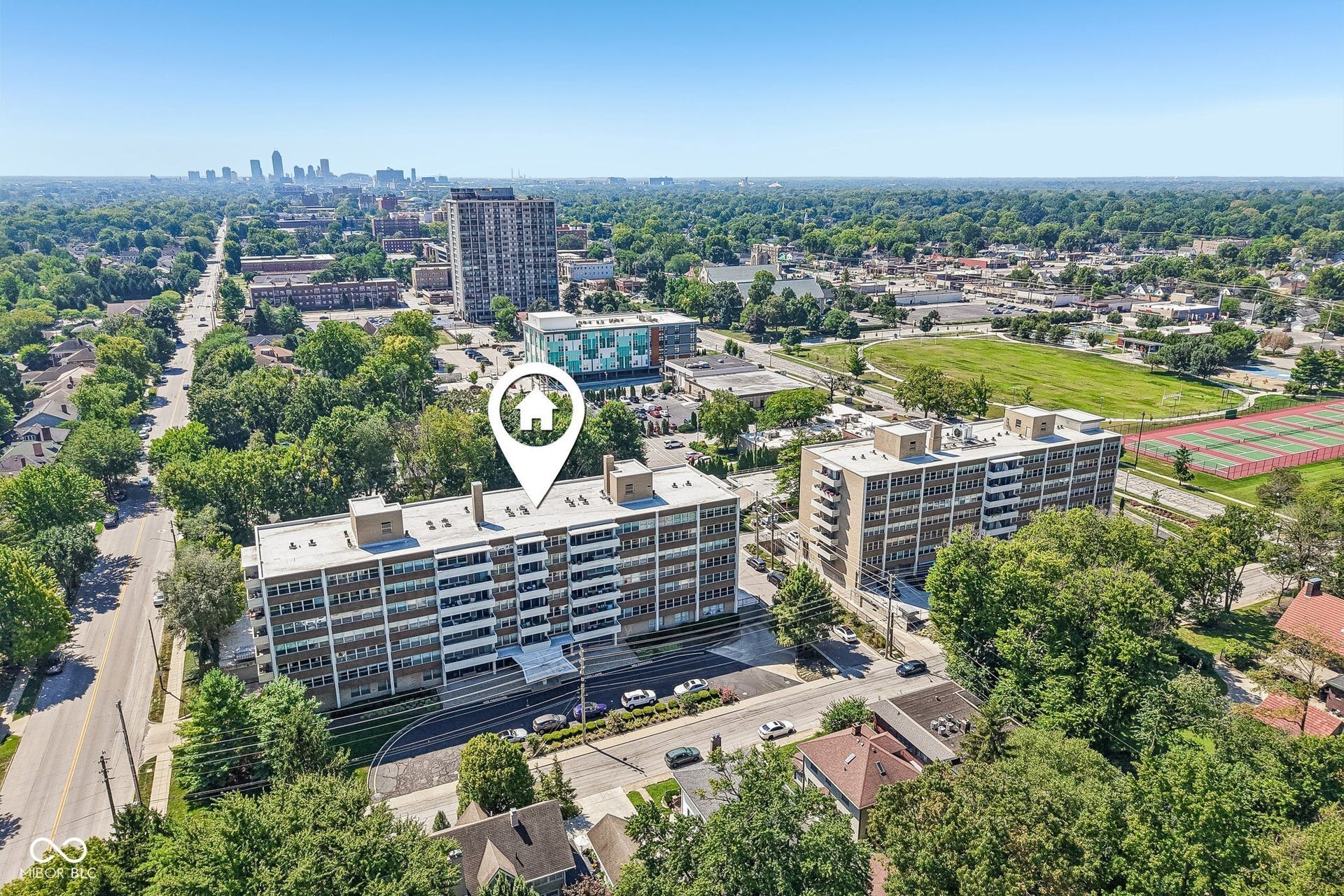 Multi-story residential building with multiple balconies, surrounded by mature trees, with a distant urban skyline