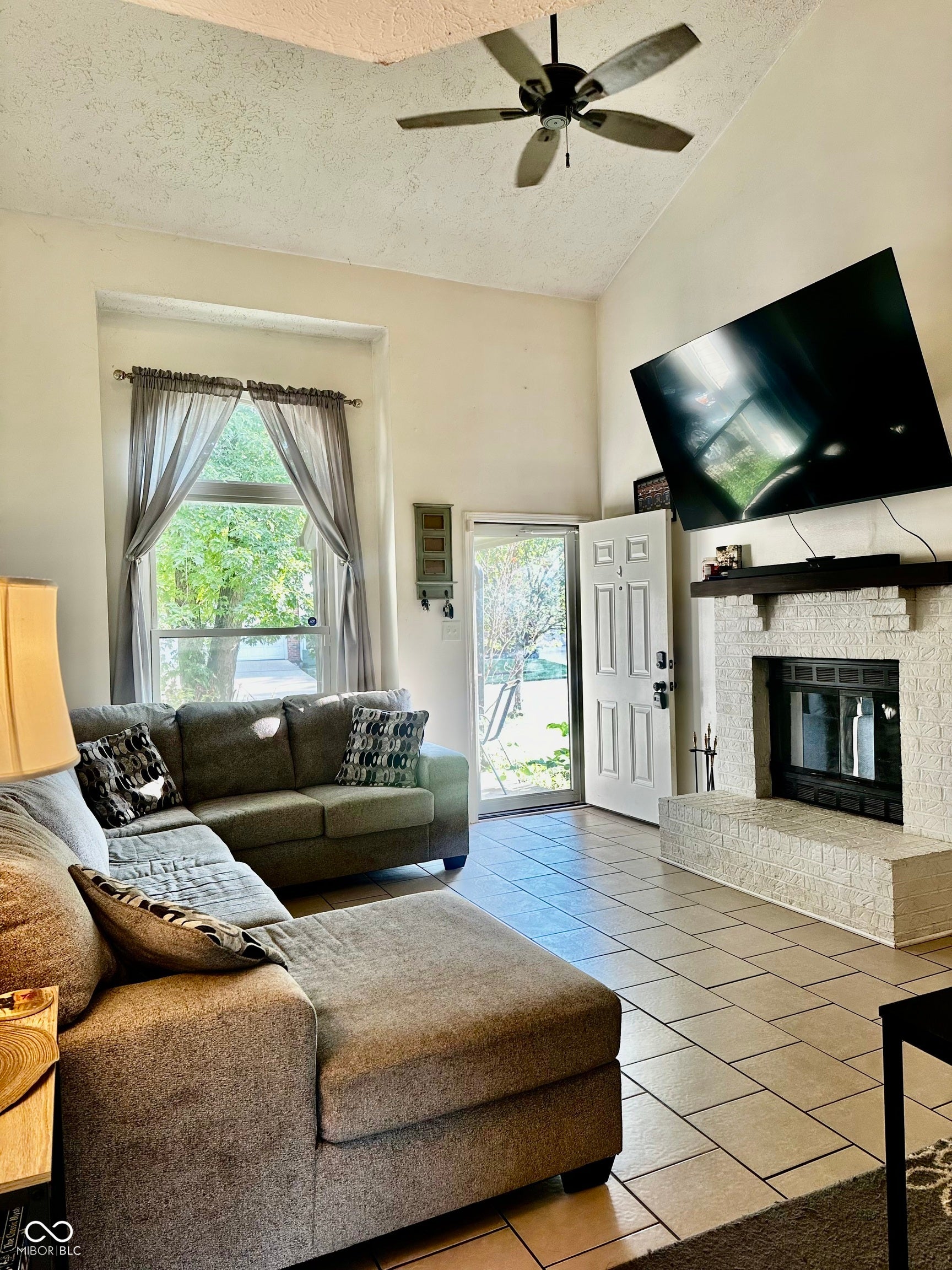 Vaulted ceiling with ceiling fan, tile flooring, white brick fireplace with dark wood mantel, and a sliding glass door