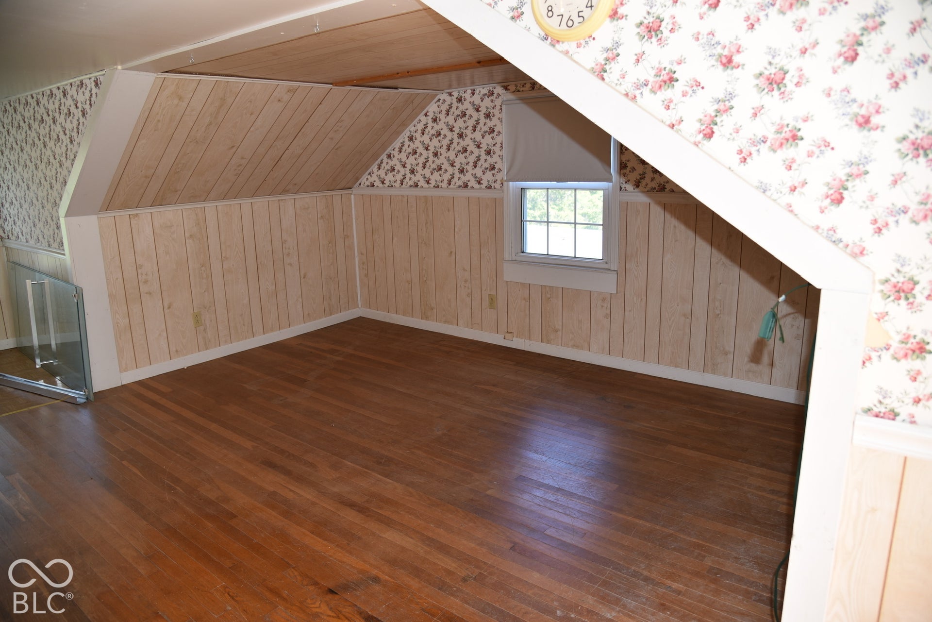 Hardwood flooring with natural finish, paneling with vertical wood-finish planks, window with white trim, and a glass-paneled barrier