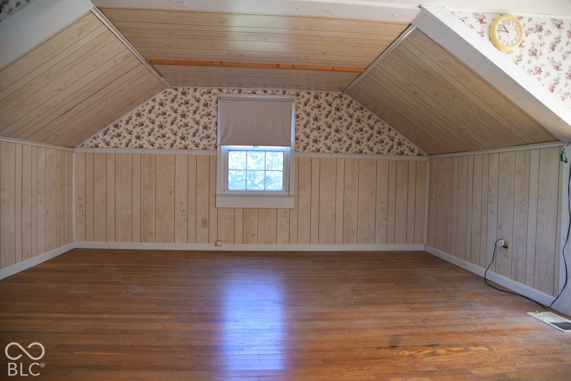 Hardwood flooring, wood-panel accent walls, sloped wood-finish ceiling, and a window with a roller shade